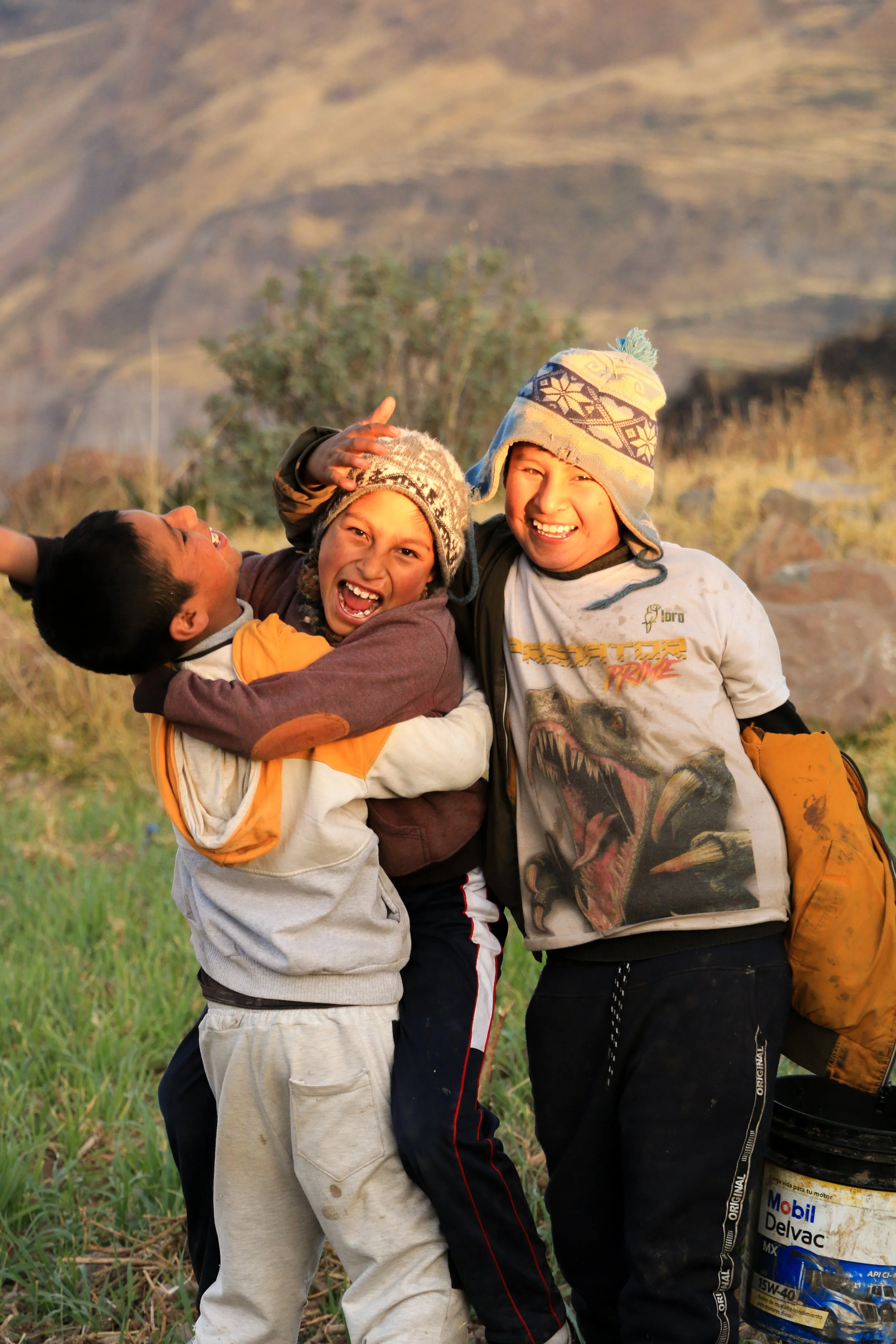 Three children smiling and hugging outdoors, wearing warm clothing, with a mountainous landscape in the background.
