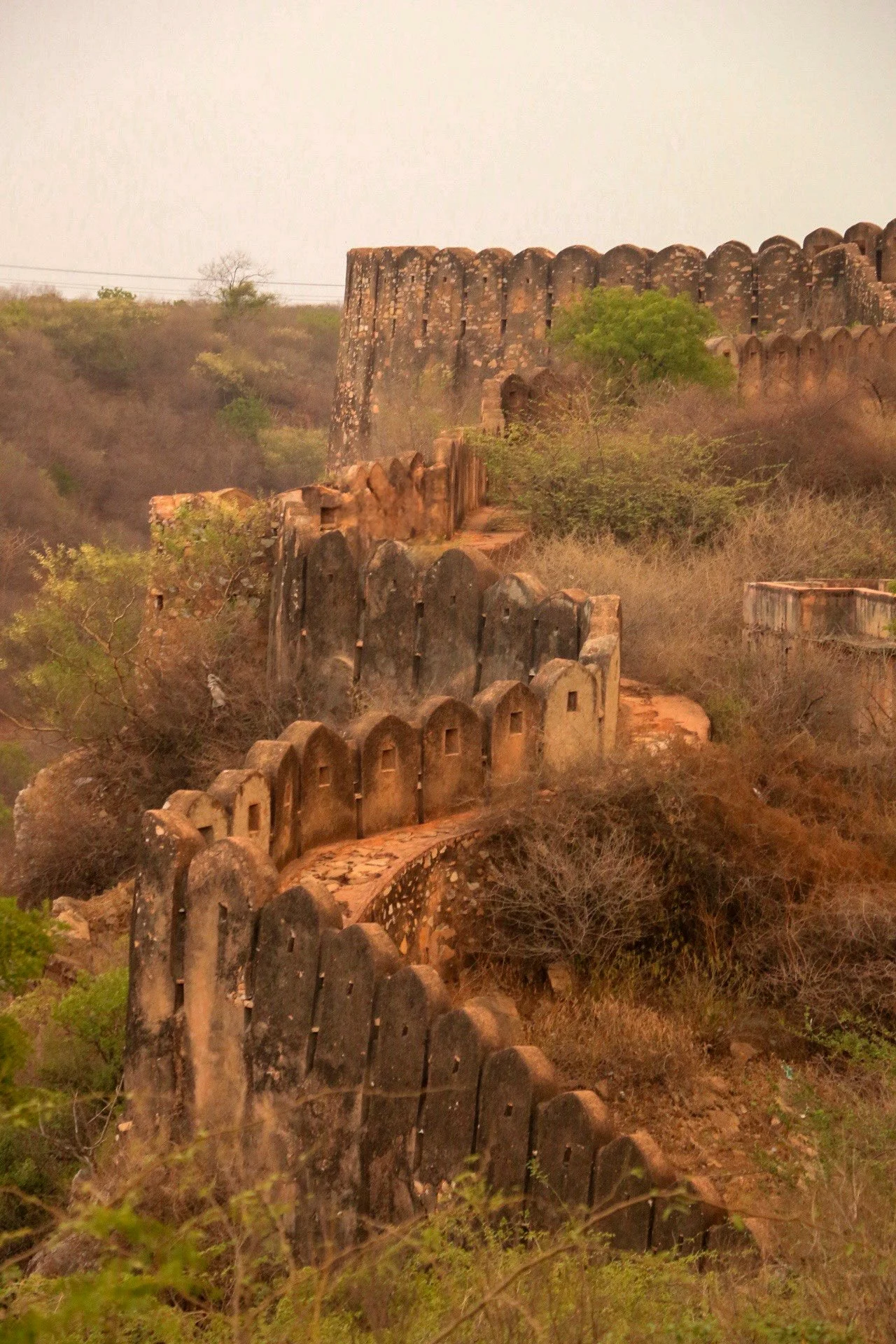Ancient stone fortification wall winding over a hilly landscape, surrounded by sparse bushes and trees, under a hazy sky.