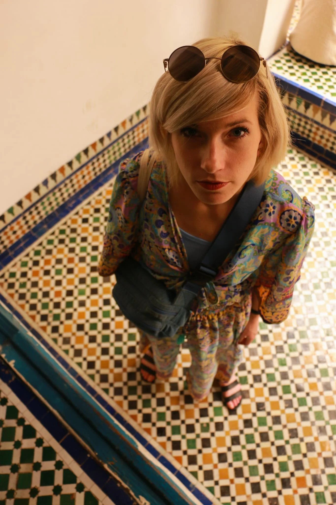 A young woman with blonde hair, wearing sunglasses on her head, standing on a colorful mosaic tile floor and looking up at the camera.