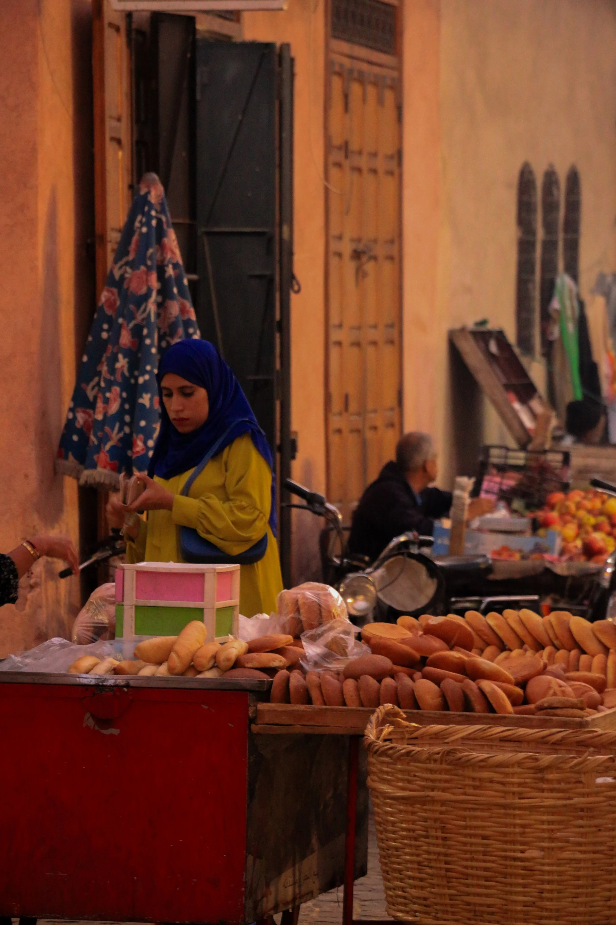 A street market scene with a woman in a blue hijab and yellow jacket standing near a table with bread, in front of a peach-colored wall. There is a colorful umbrella beside her and a woman sitting on a motorbike in the background.