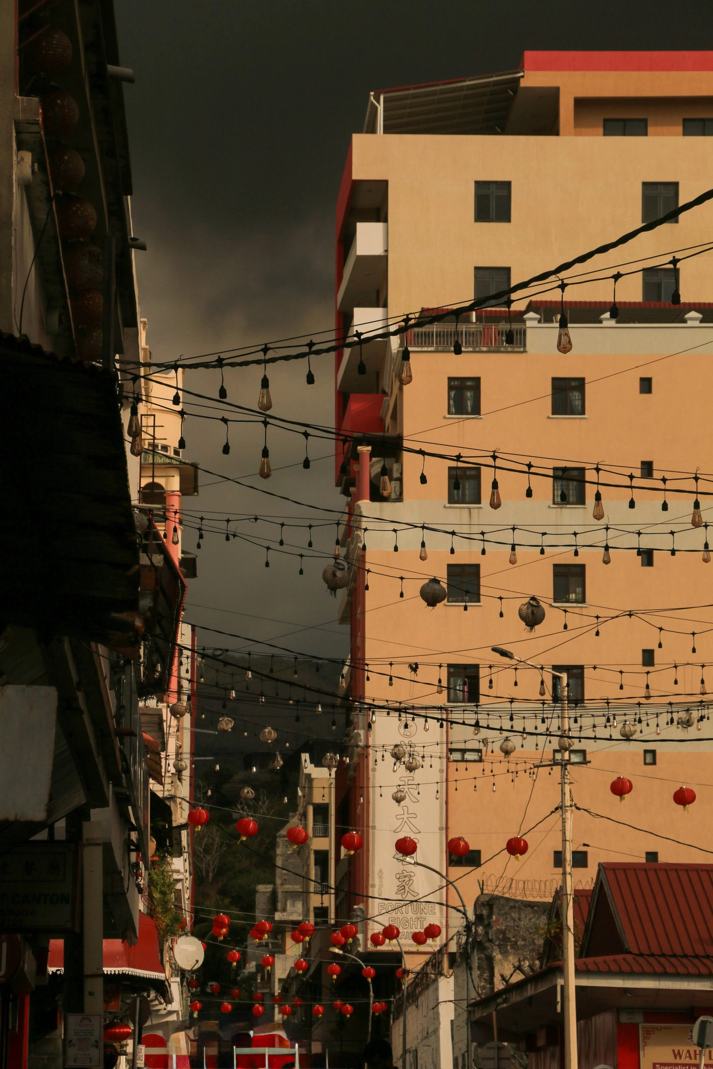 A city street with hanging string lights and red lanterns, tall buildings, and a dark stormy sky in the background.