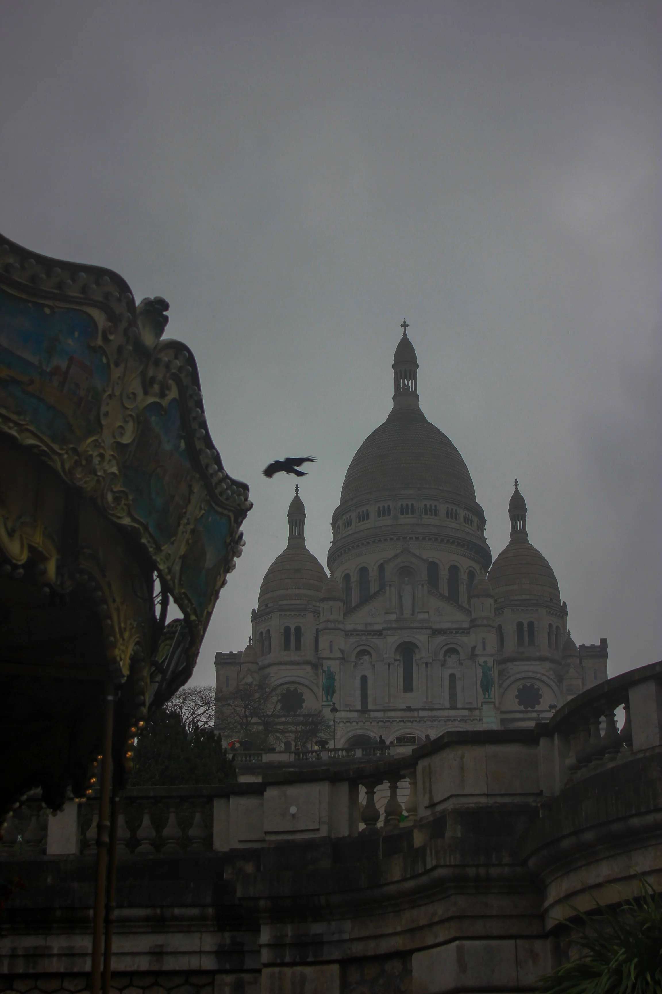 The Sacré-Cœur Basilica in Paris, France, with a cloudy sky and a flying bird, partially visible ornate carousel on the left, and stone railing in the foreground.