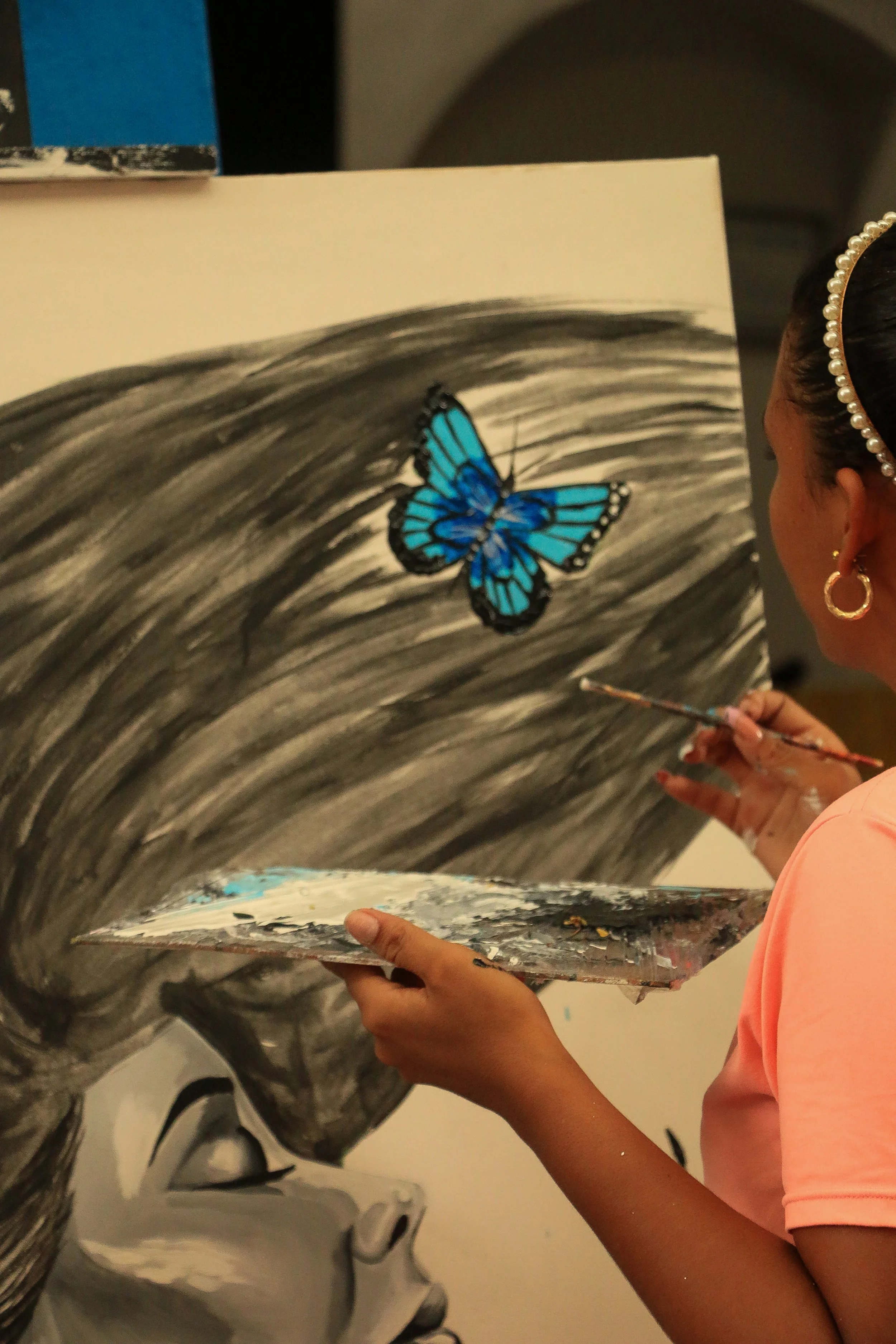 An artist is painting a black and white portrait of a face on a canvas, with a blue butterfly as part of the artwork.