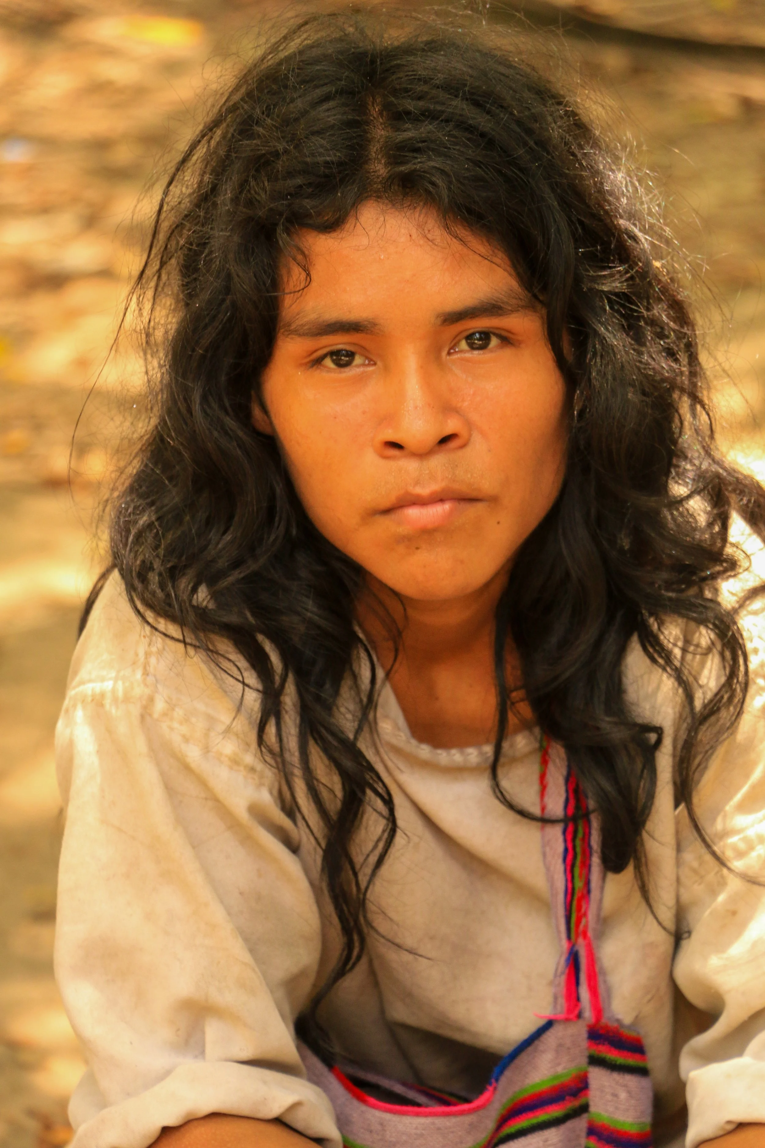 A young woman with wavy black hair and tan skin looking directly at the camera with a serious expression, wearing a cream-colored shirt with colorful embroidery, outdoors with blurred autumn leaves in the background.