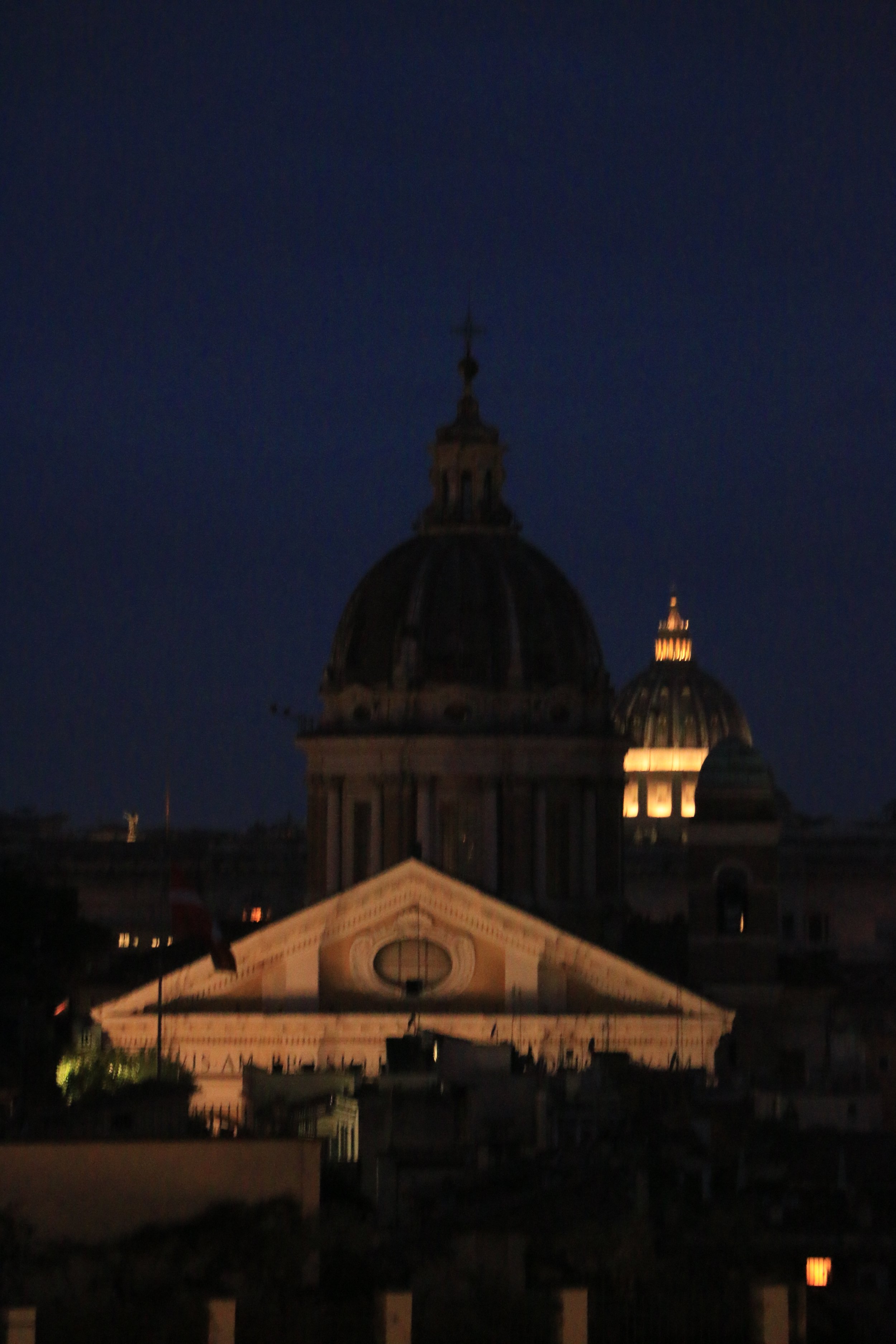 Nighttime view of a historic building with a large domed roof and illuminated windows, likely a church or cathedral, with a dark sky in the background.