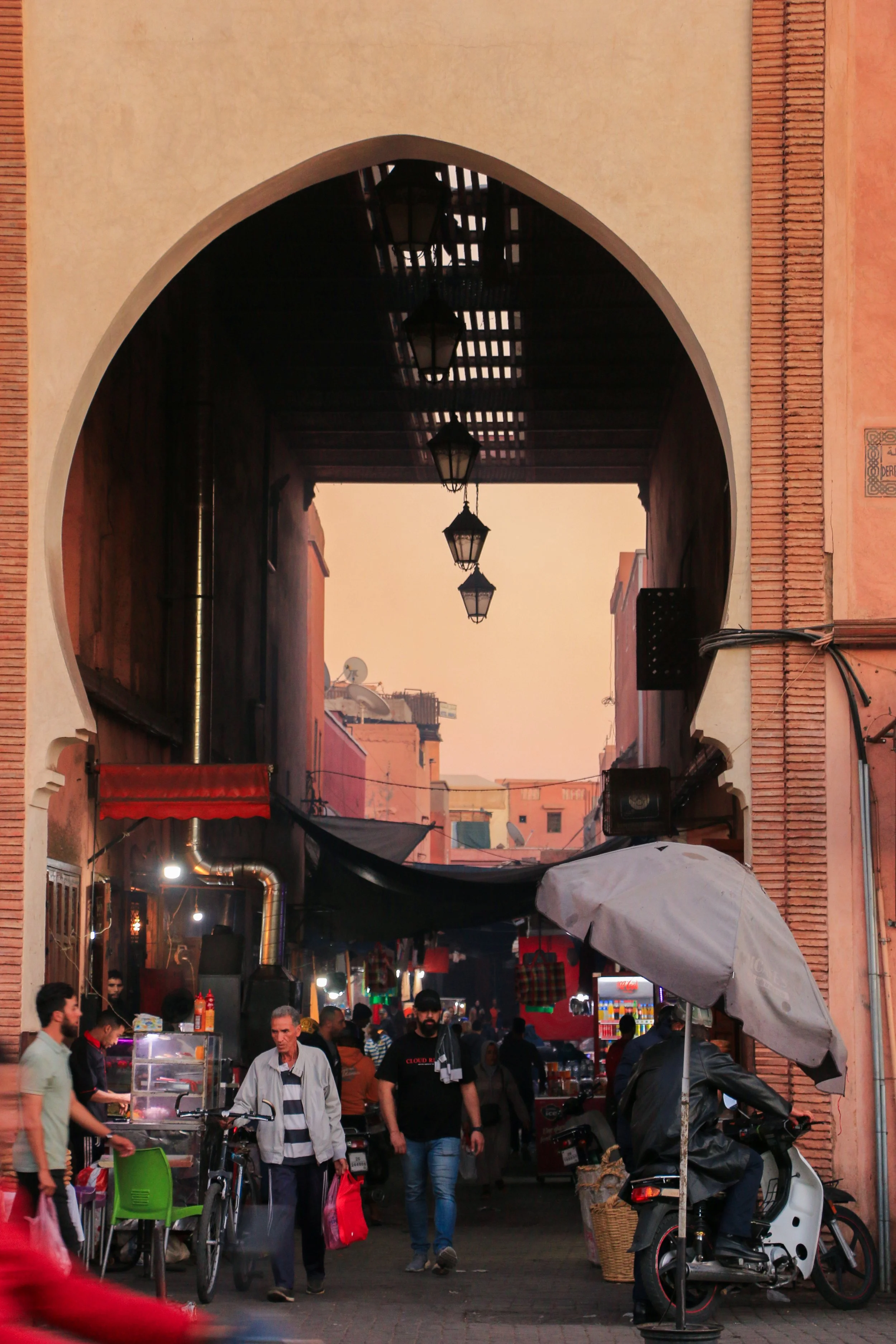 A bustling market street in Morocco with an archway and hanging lanterns, featuring people walking, a vendor on a motorcycle, and colorful storefronts.