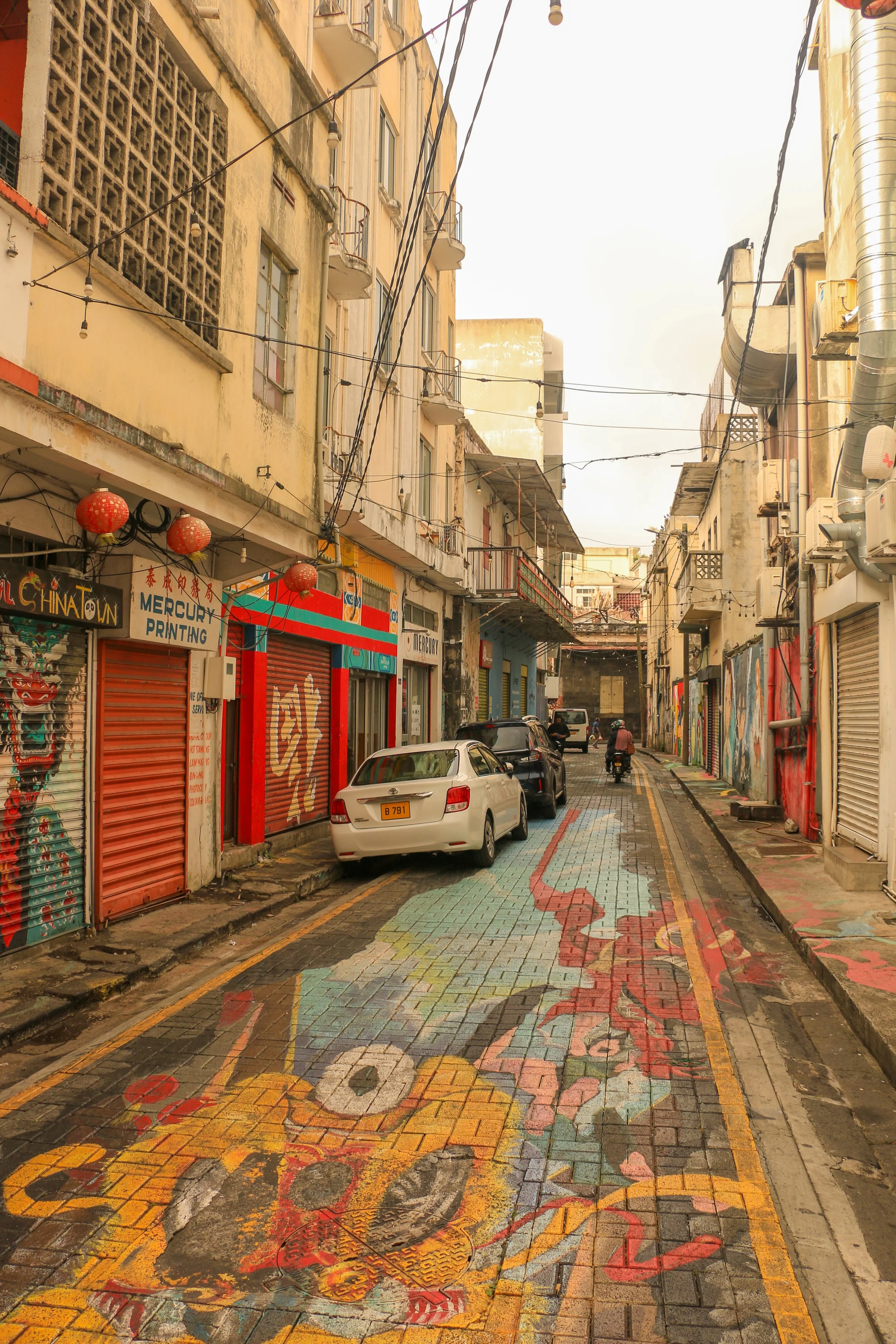 A narrow urban street with colorful murals painted on the pavement and old buildings lining both sides. Several cars and motorcycles are parked or moving along the street, which is decorated with hanging lanterns and overhead wires.