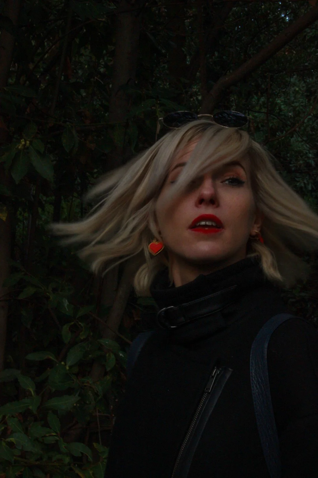 A woman with blonde hair blowing in the wind, wearing red lipstick, red earrings, and black clothing, standing in a dark forested area.