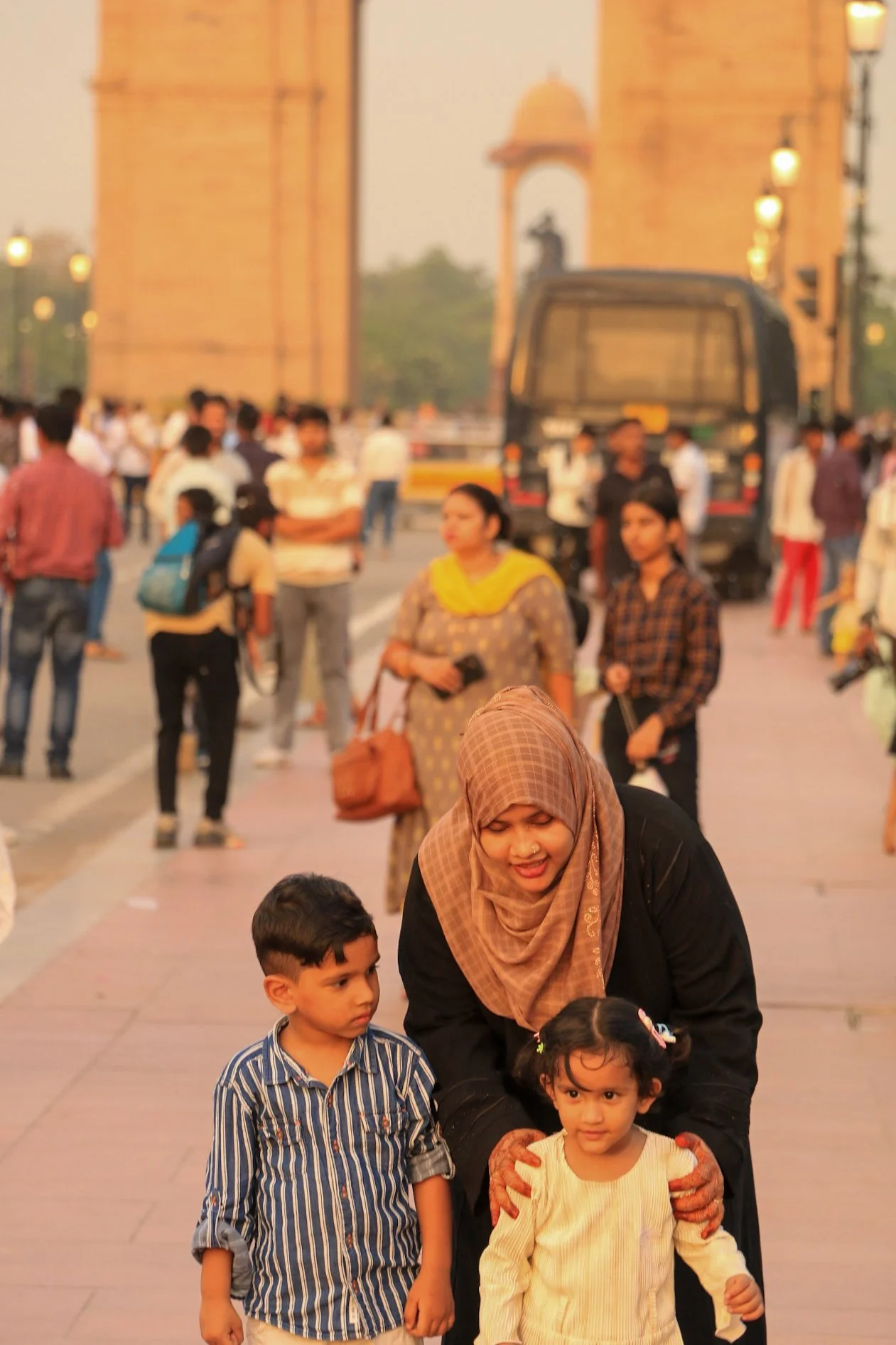 A woman with a headscarf and two children, a boy and a girl, on a busy city street near India Gate with many people and a black vehicle in the background.