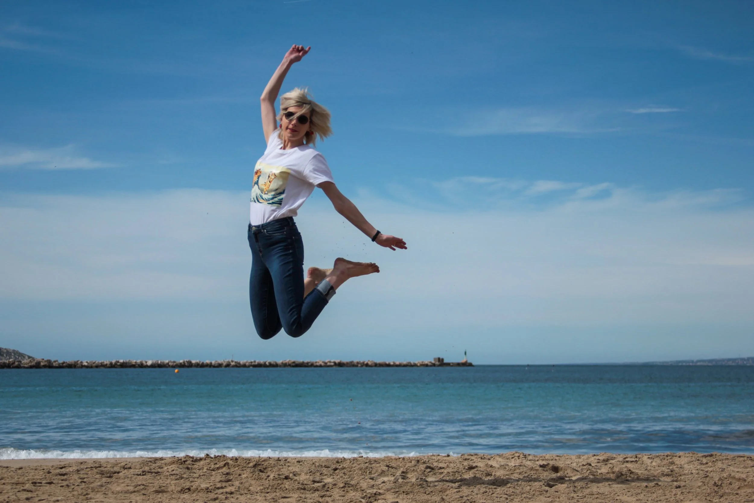 A woman with blond hair, wearing sunglasses, a t-shirt with a graphic of a boat, and jeans, is jumping on a sandy beach near the ocean, with a clear blue sky above and a breakwater with a lighthouse in the distance.