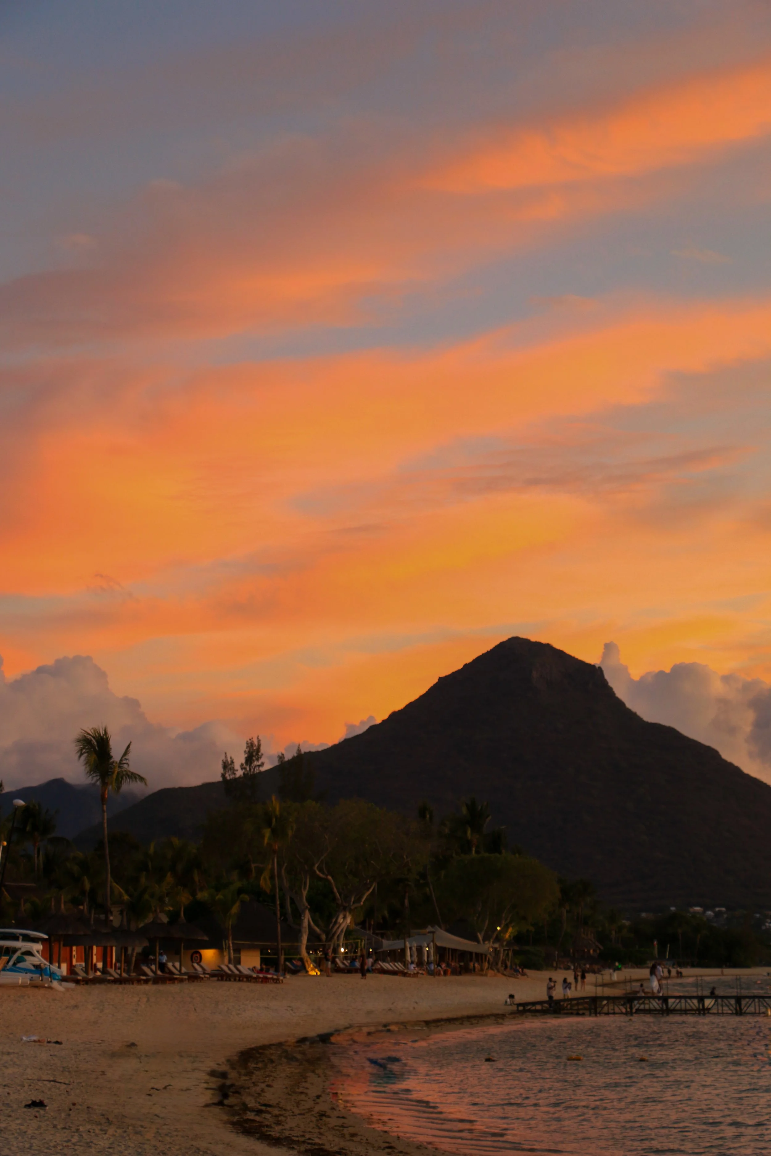A beach scene during sunset with colorful sky, mountain in the background, and people walking along the shore.
