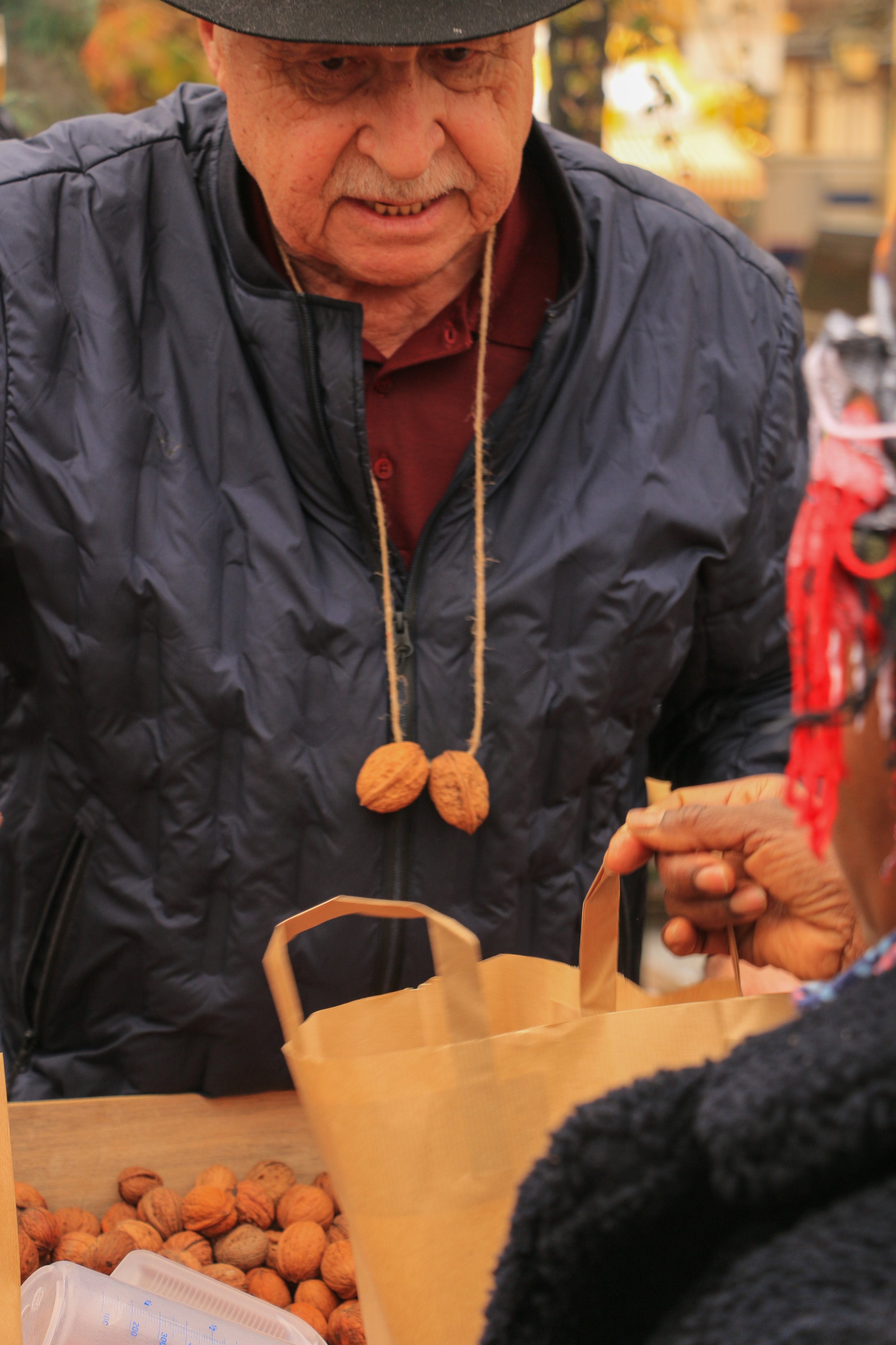 An elderly man wearing a black jacket, maroon shirt, and black hat is selling or giving out walnuts at an outdoor market. The image shows a basket of walnuts in front of him and a person on the right receiving some walnuts.
