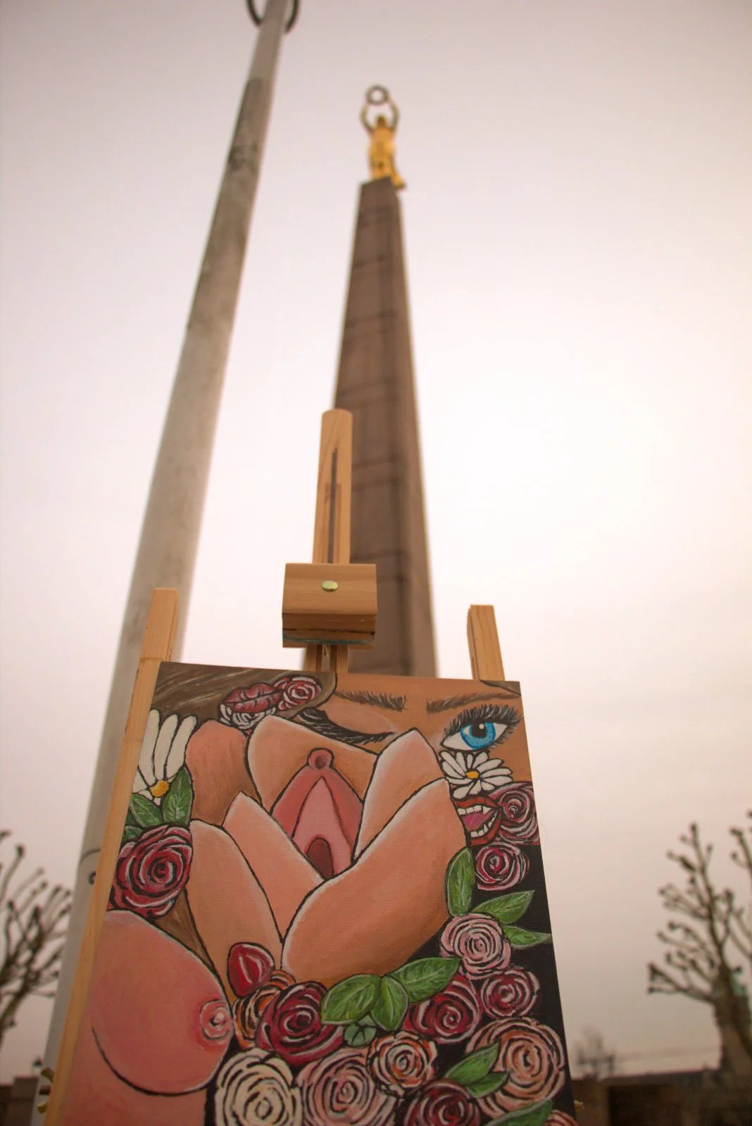 A painting of a woman’s face, with one eye visible, surrounded by roses, with an umbrella, in front of a tall monument topped with a golden figure, and a streetlight.
