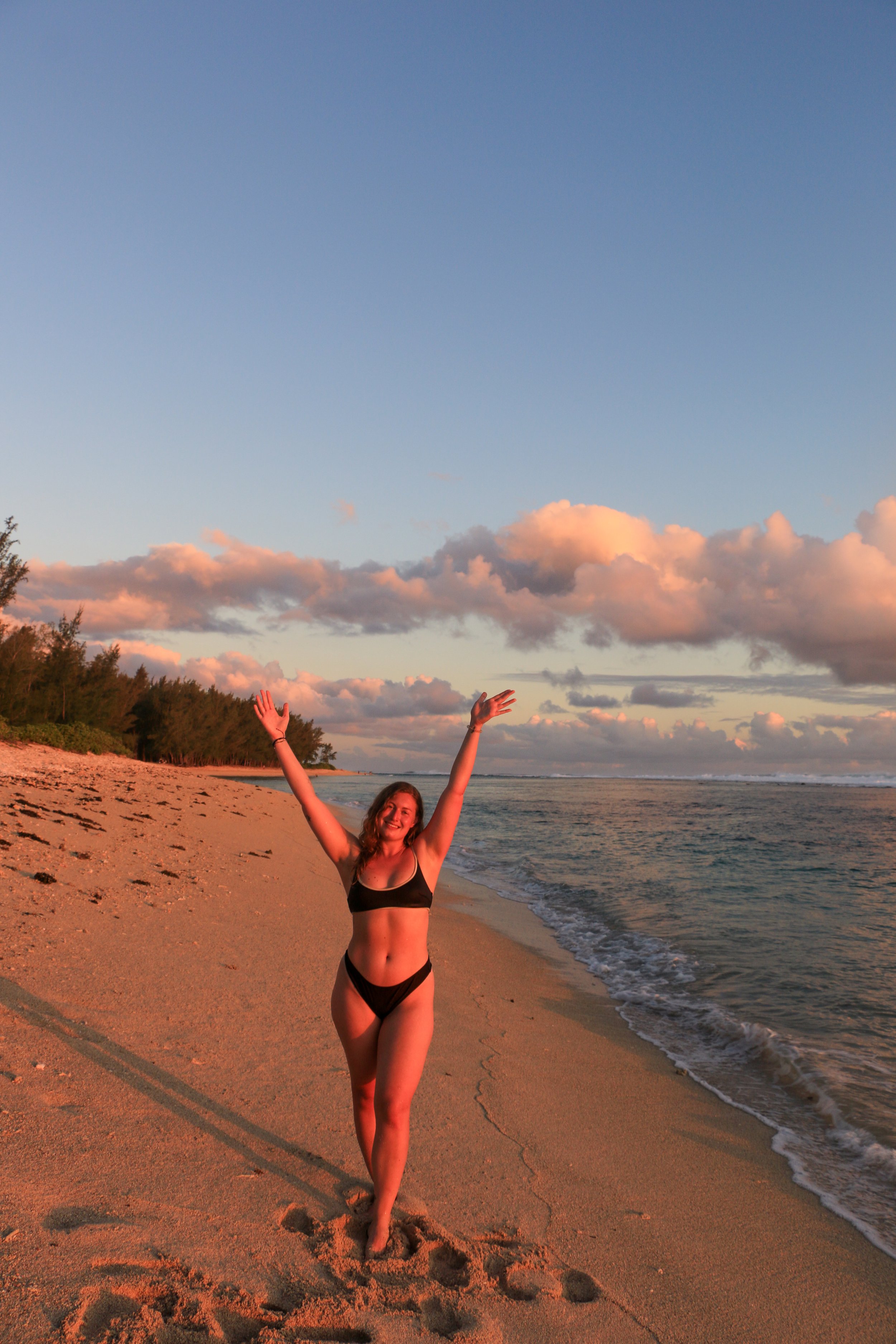 Woman in black bikini celebrating on a sandy beach during sunset with clouds in the sky.