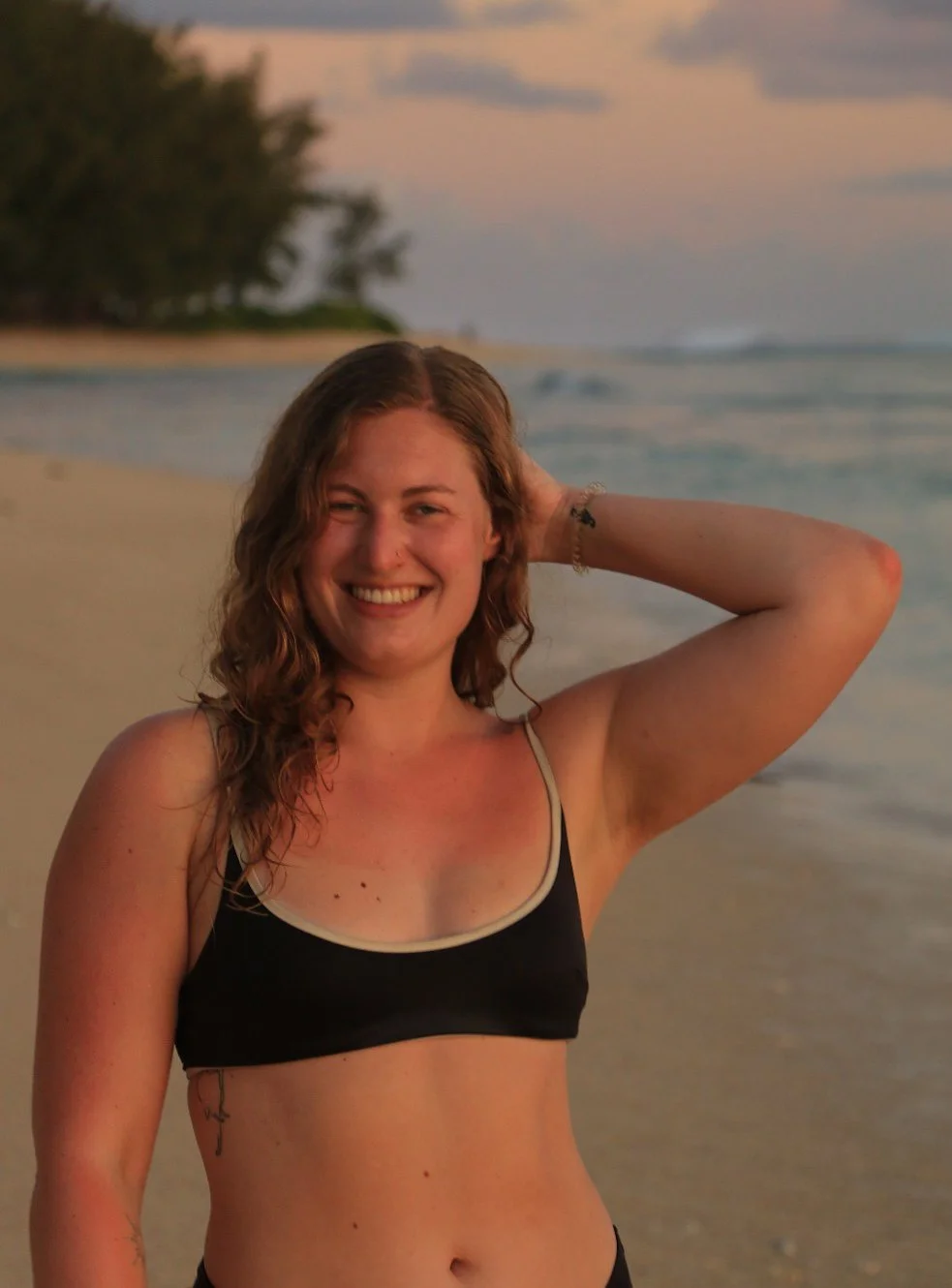 Young woman with wavy reddish hair smiling on a beach at sunset, wearing a black bikini top, with one hand behind her head, and trees in the background.