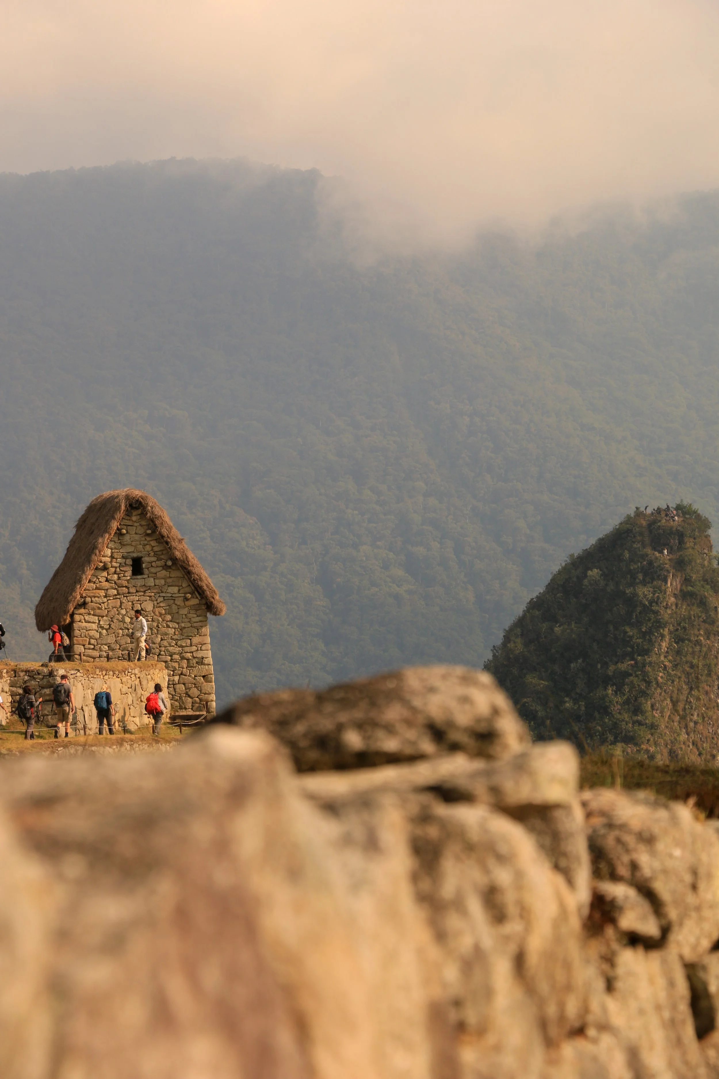 A stone building with a thatched roof in a mountain landscape with several hikers in the foreground and green hills in the background.