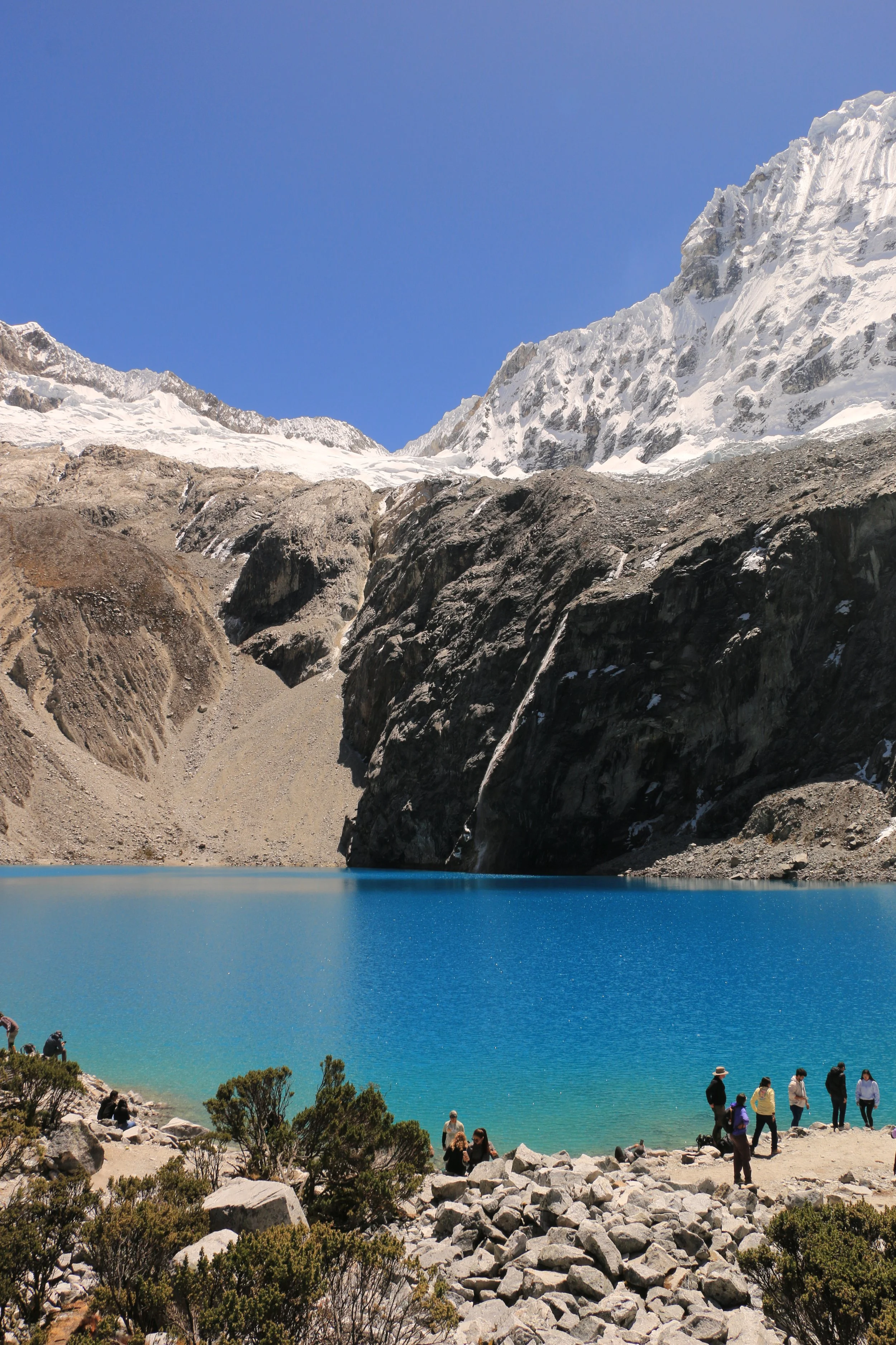 Tourists gathered near a turquoise lake surrounded by rocky terrain and snow-capped mountains in the background.