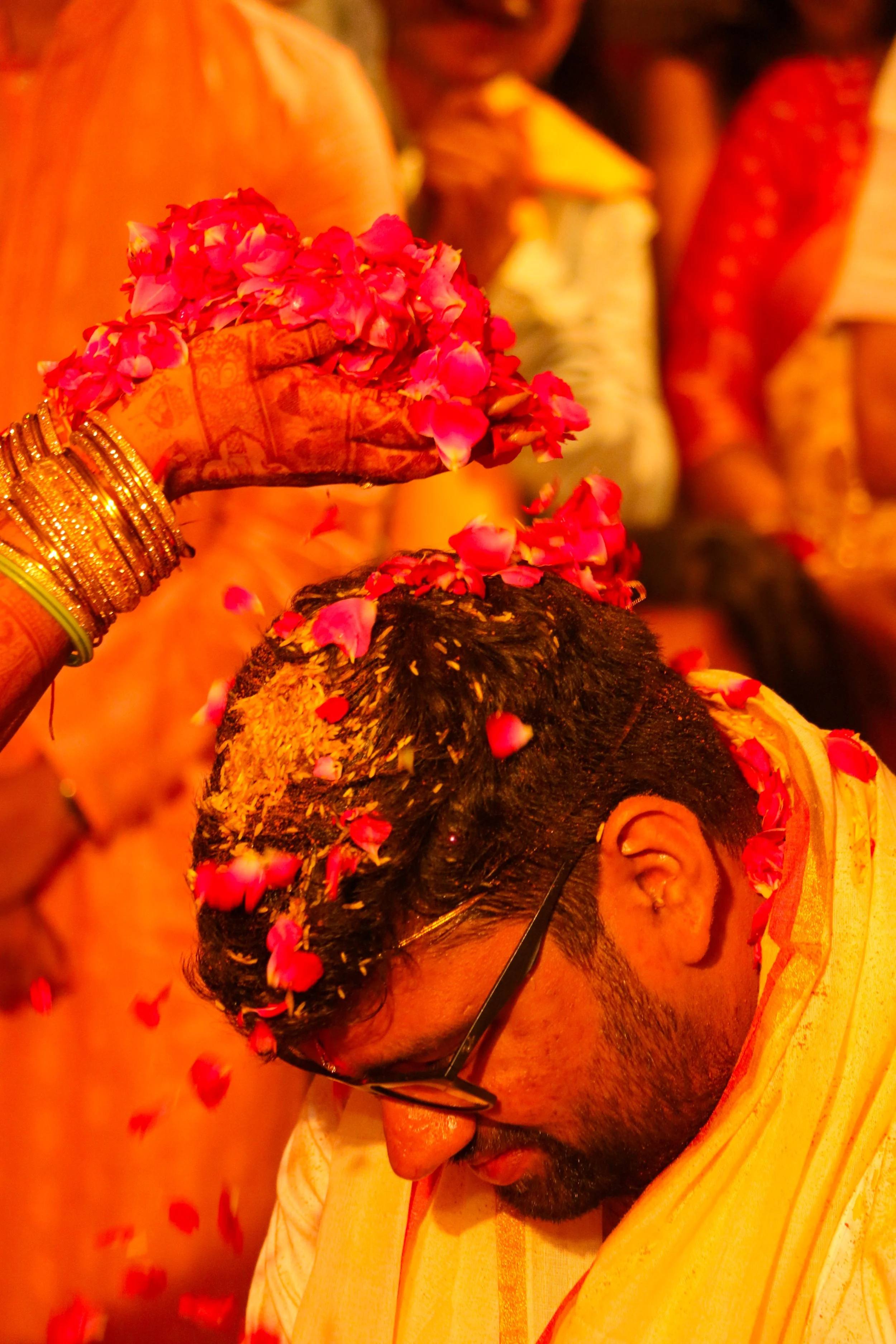 A person with dark hair and glasses participates in a traditional ceremony, with pink flower petals being sprinkled on their head, surrounded by others dressed in colorful clothing.