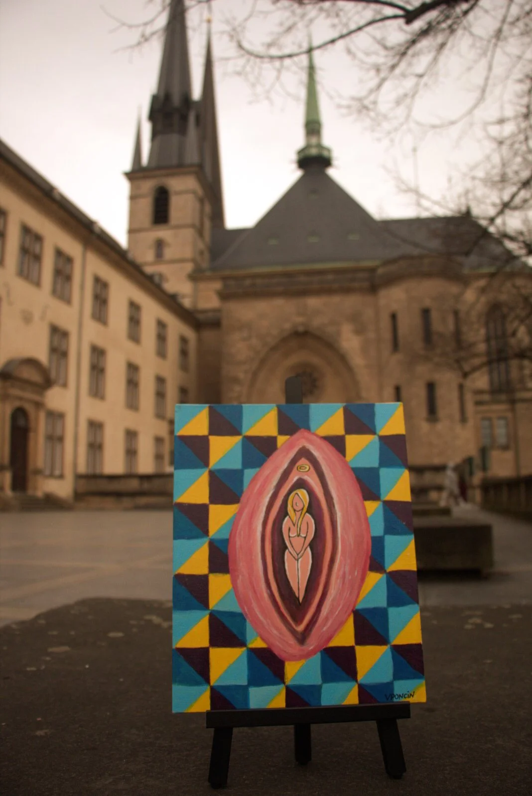 A small painting showing a stylized figure with a halo, inside an eye-shaped outline. The painting background features a geometric pattern of blue, yellow, and brown triangles. The painting is displayed on a small easel outdoors in front of a historical stone building with two steeples, one tall and dark and the other shorter with a green cap, and leafless trees in the background.