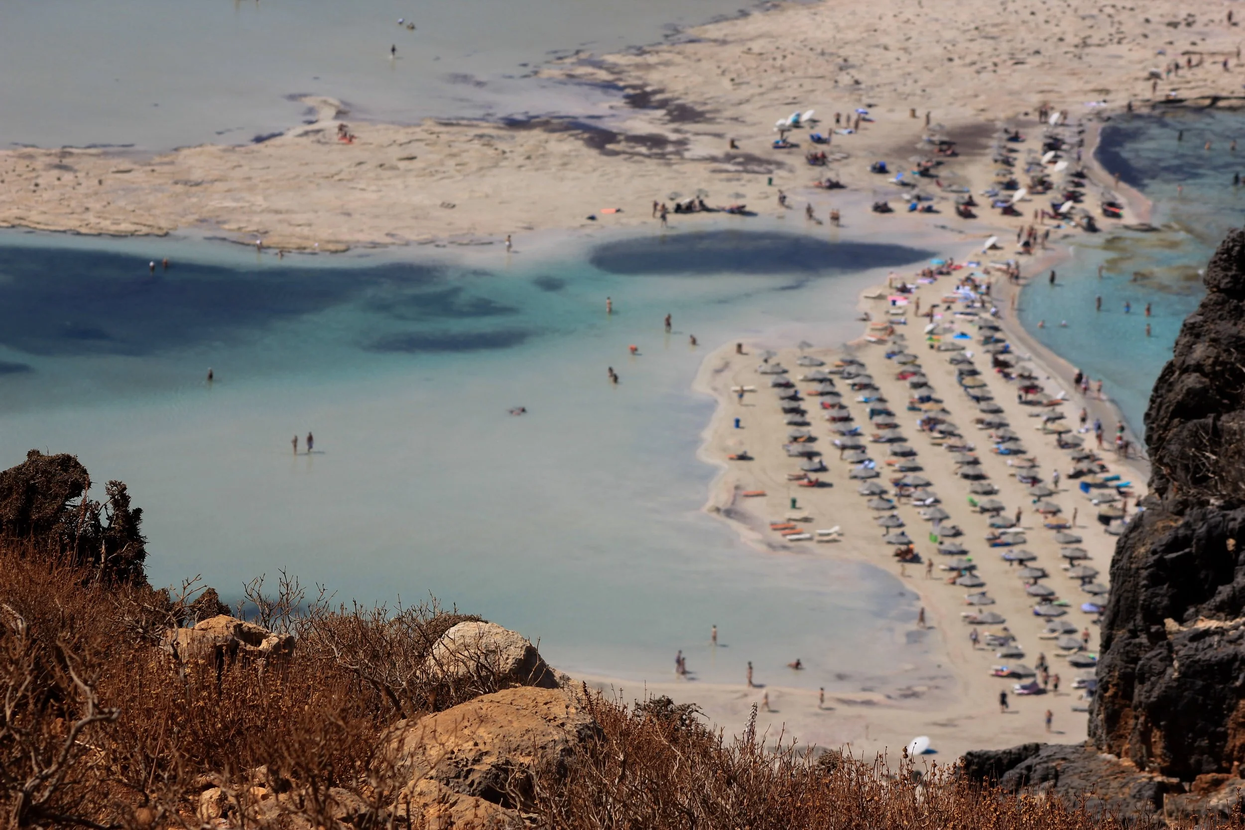 Aerial view of a crowded beach with lounge chairs and umbrellas, turquoise water, and rocky terrain in the foreground.