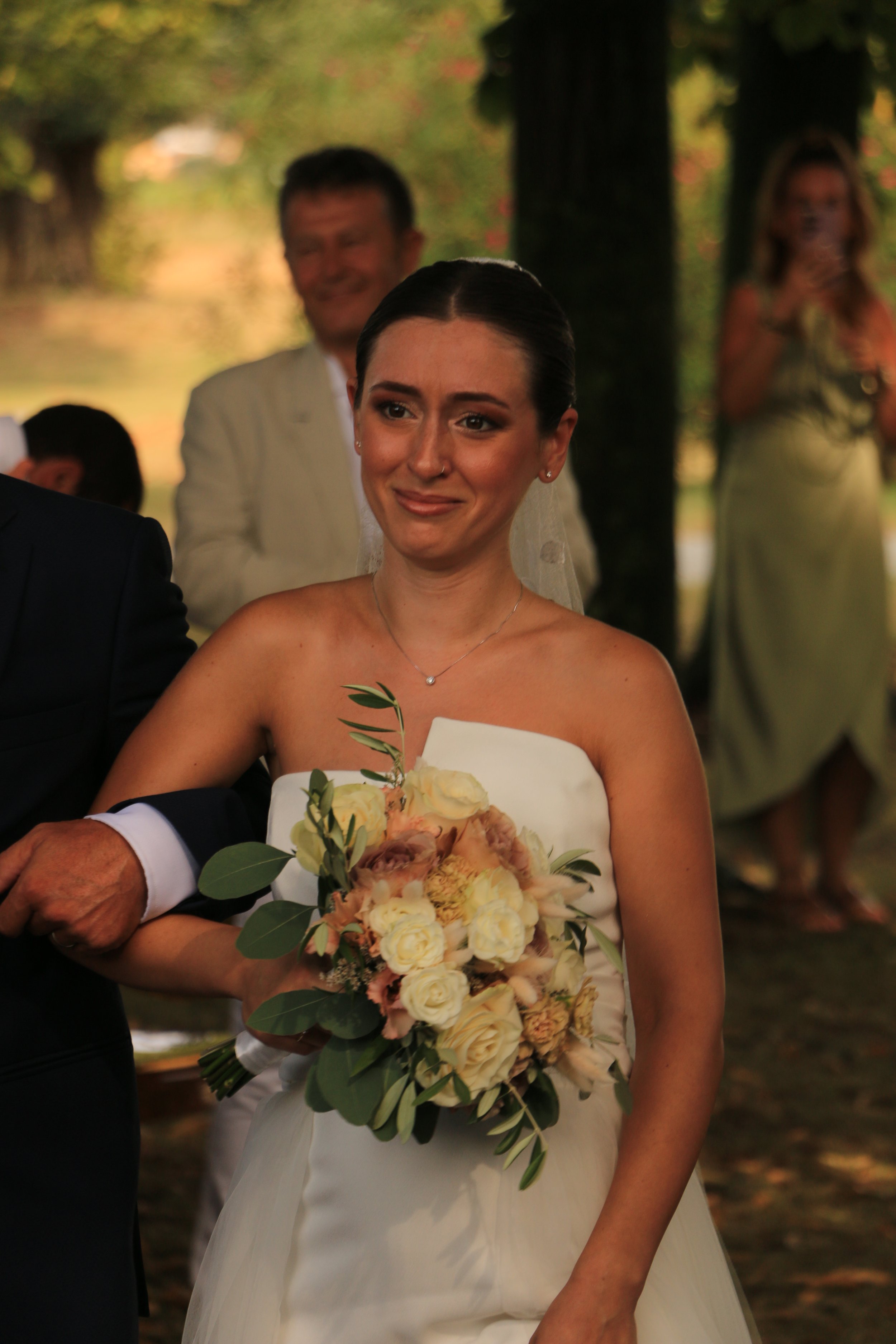 Bride in a white strapless wedding dress holding a bouquet of cream-colored roses and greenery during an outdoor wedding ceremony.
