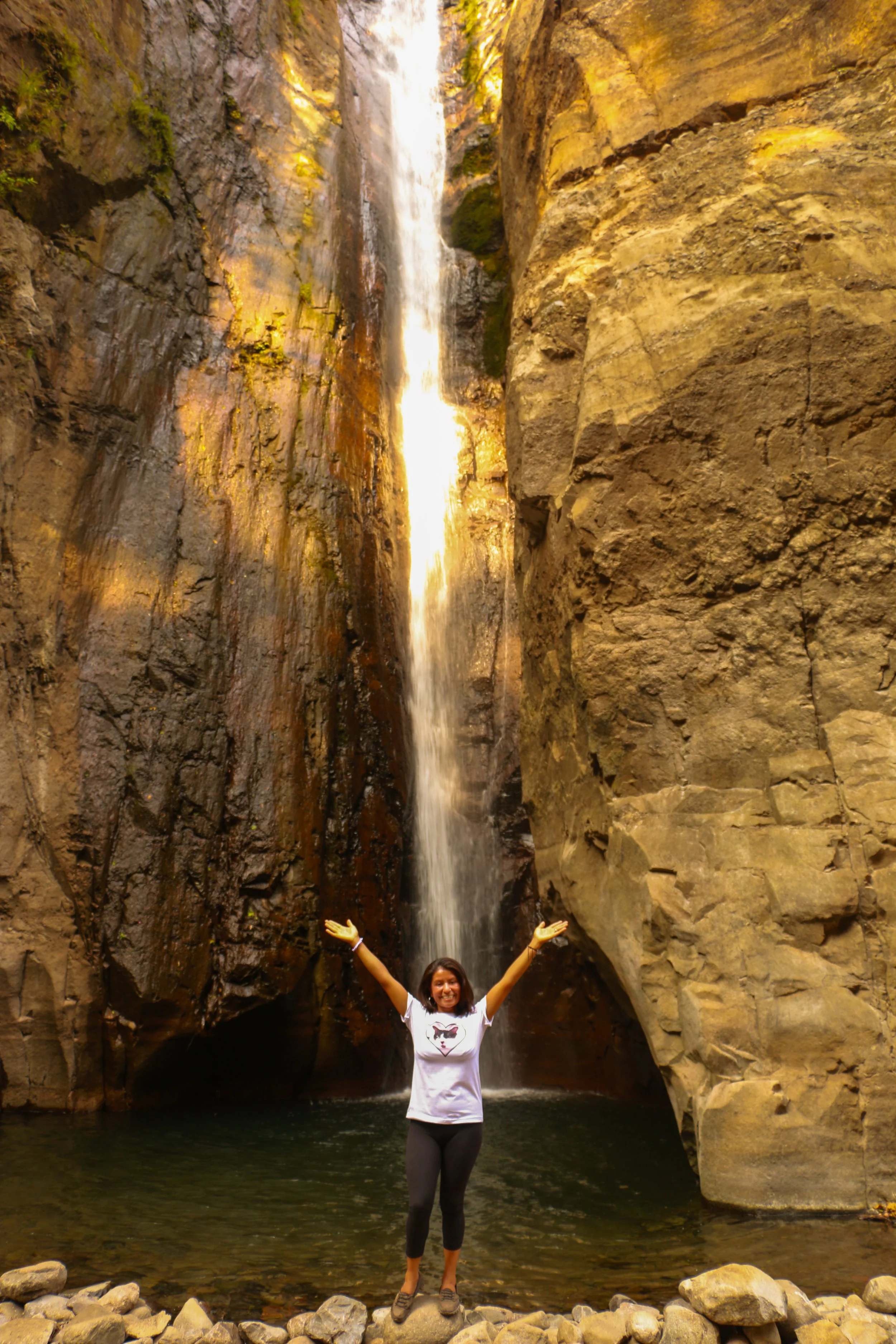 A woman standing with arms raised in front of a waterfall inside a canyon with rocky walls.