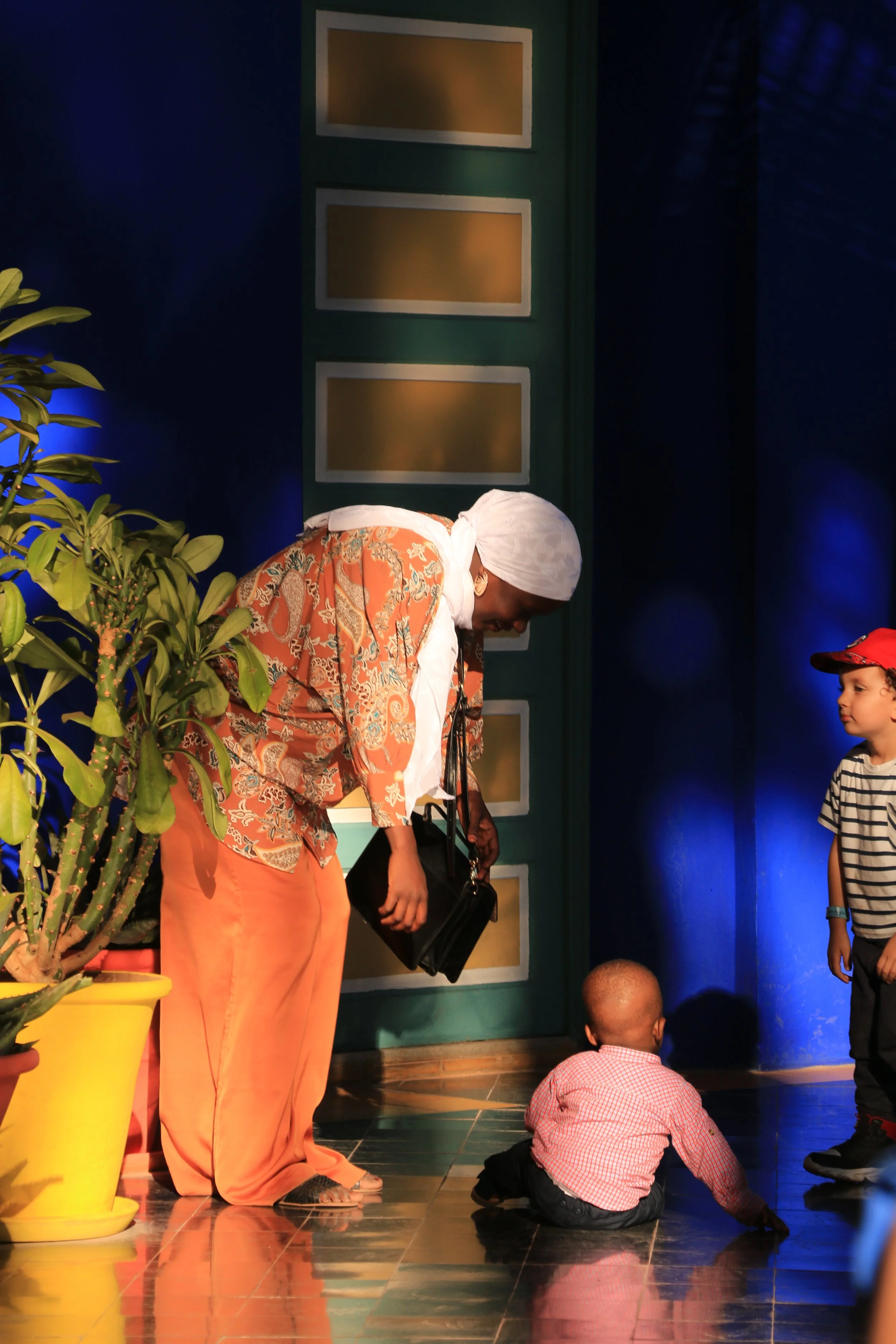 A woman with a white headscarf and orange patterned outfit bending down to speak to a seated child on the floor. Another young boy wearing a striped shirt and red cap standing nearby. Potted plant on the left side and background with colorful decor.