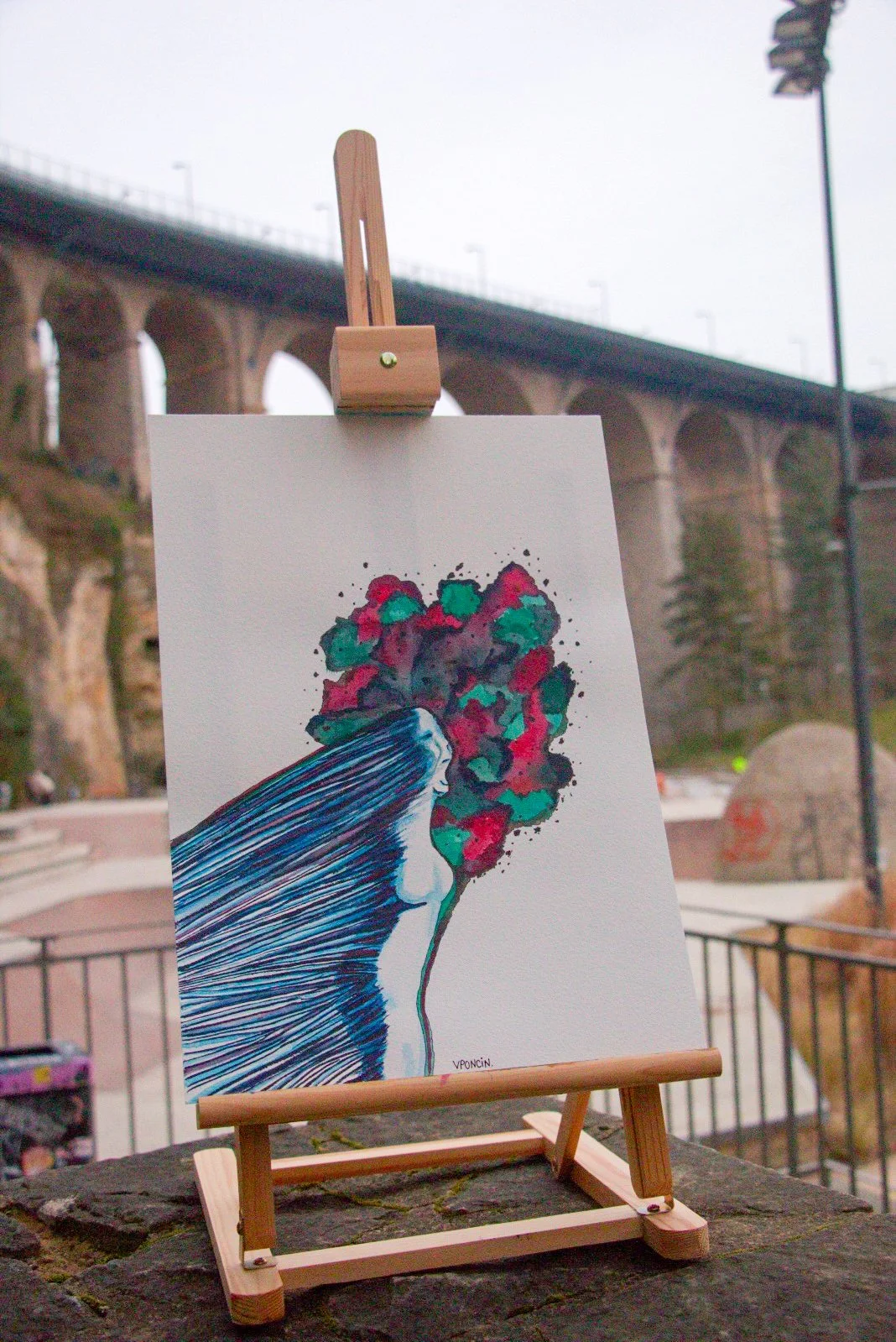 A colorful watercolor artwork of a woman with flowing blue hair, with her face turned sideways, and hair transforming into a vibrant explosion of red, green, and black colors, displayed on a small wooden easel outdoors with a bridge and trees in the background.