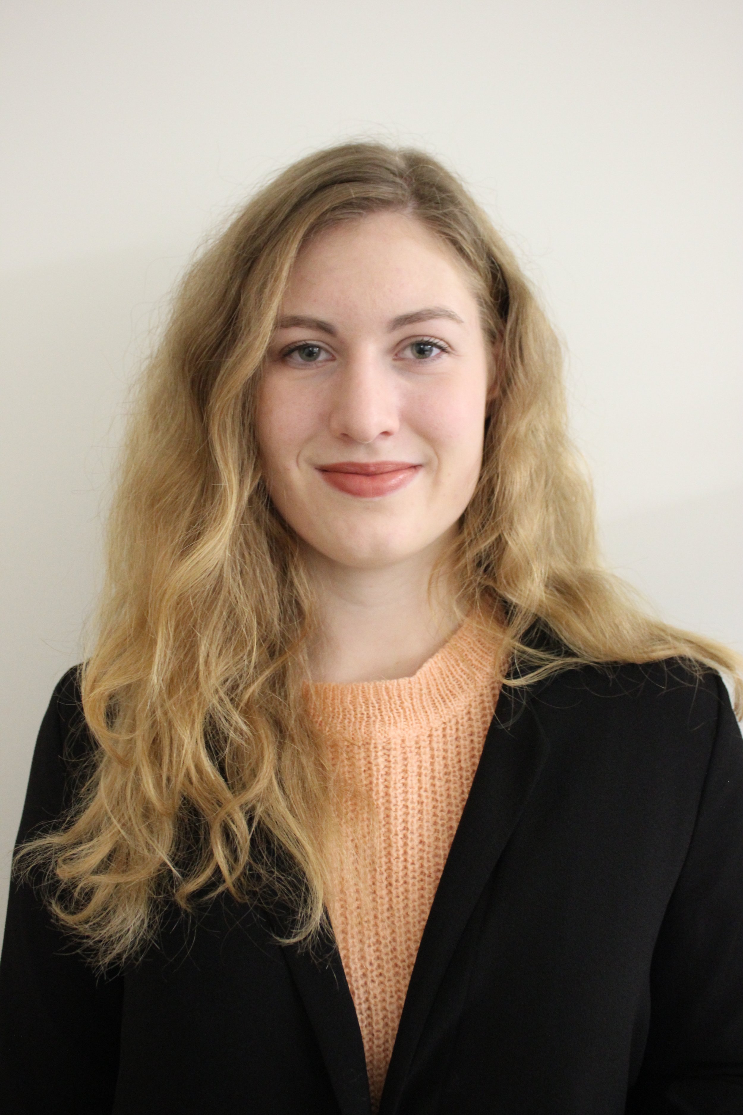 Portrait of a young woman with long, wavy blonde hair, wearing a black blazer over a peach-colored knit top, standing against a plain, light-colored background.