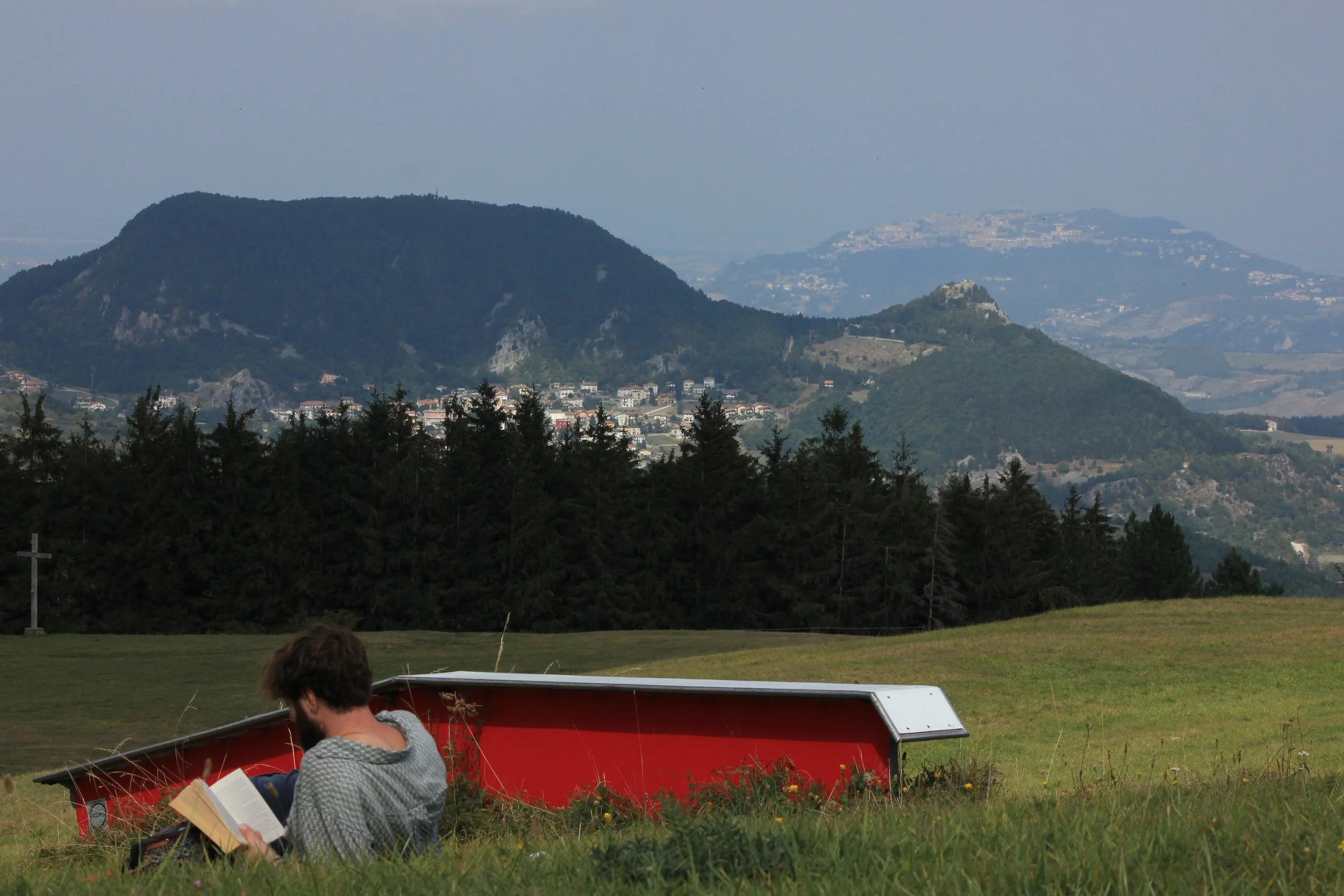 A man sitting in a grassy field reading a book, with an upside-down red boat beside him and a mountain landscape in the background.