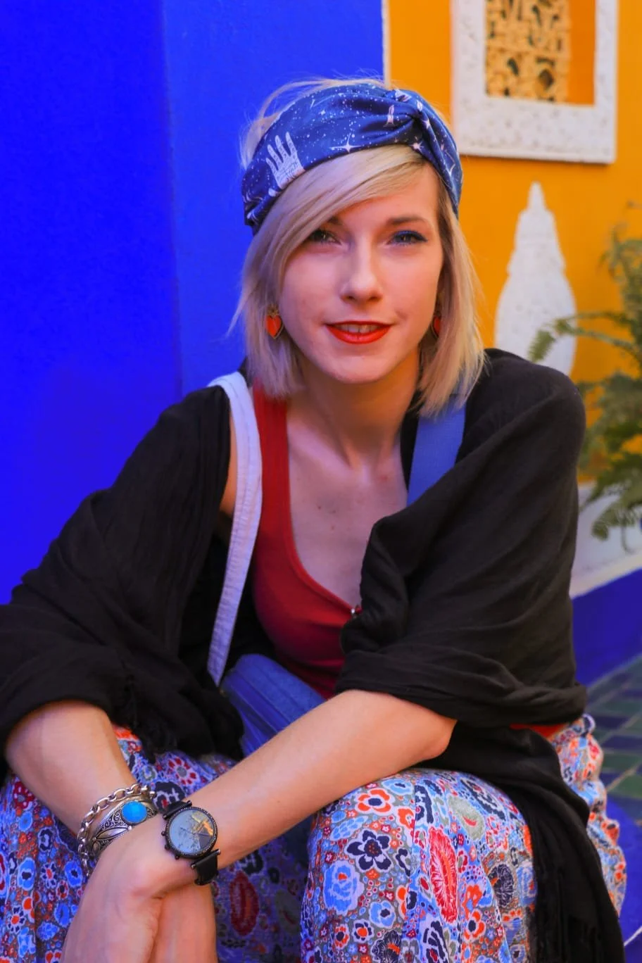 A young woman with blonde hair wearing a blue bandana, red lipstick, and colorful clothing, sitting indoors against vibrant orange and blue painted walls, with decorative art and a plant in the background.