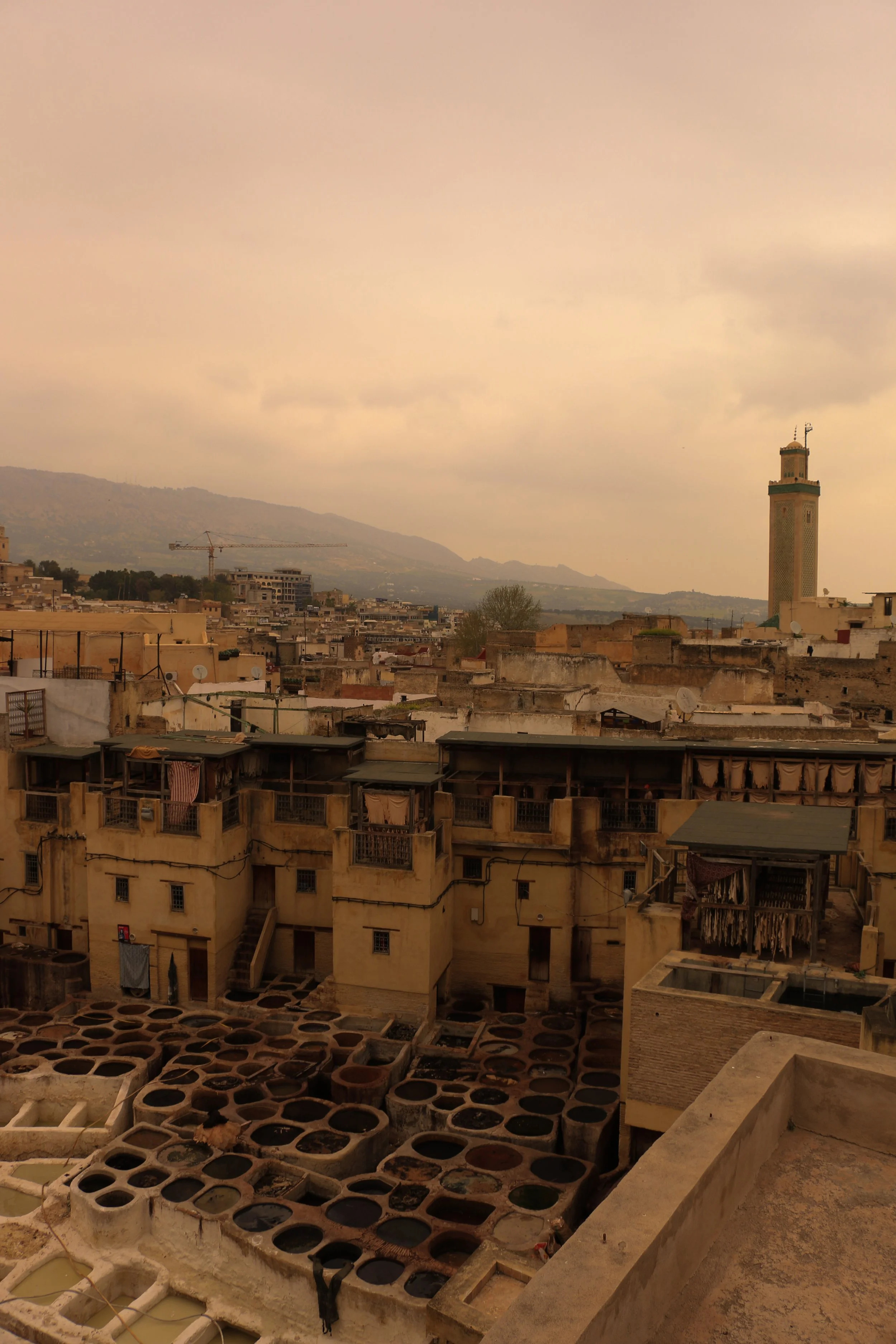 A cityscape with traditional beige buildings, a prominent minaret, and dye pits in the foreground, possibly in Marrakech, Morocco.