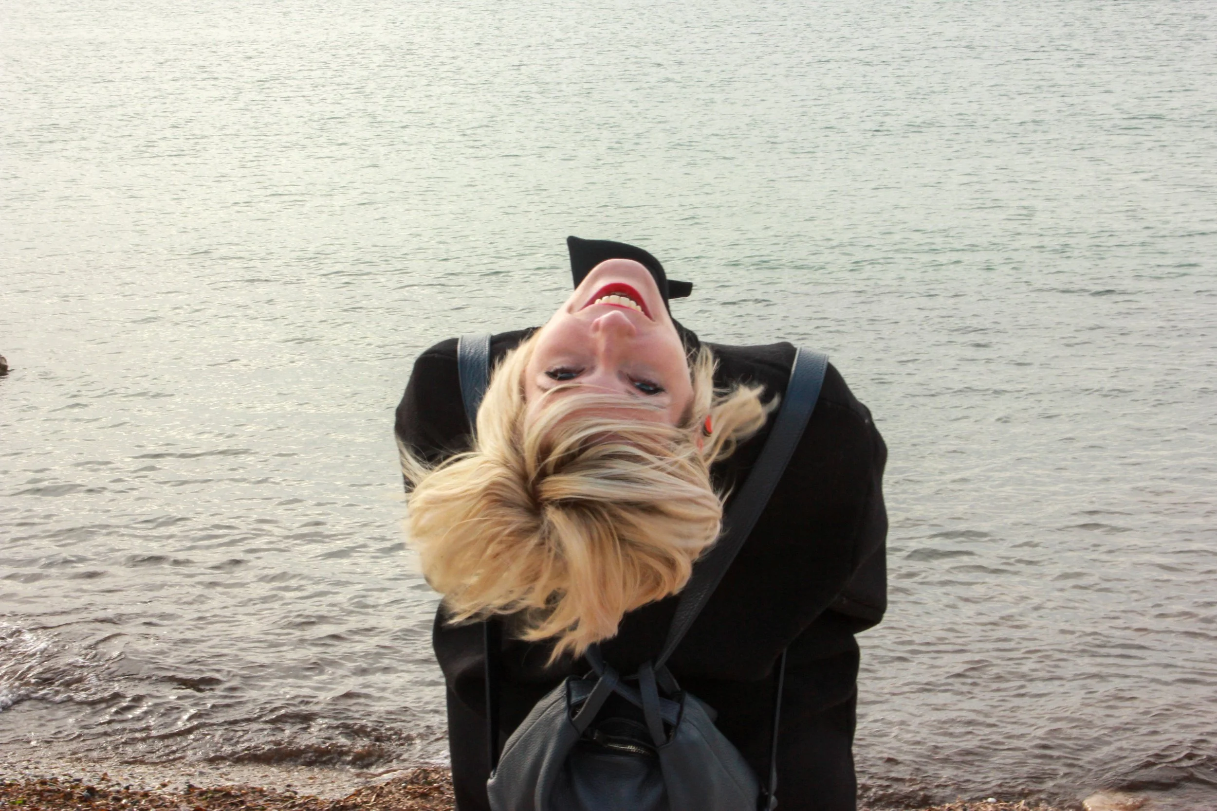 Woman with blonde hair leaning back by a body of water, smiling with her head tilted upside down.
