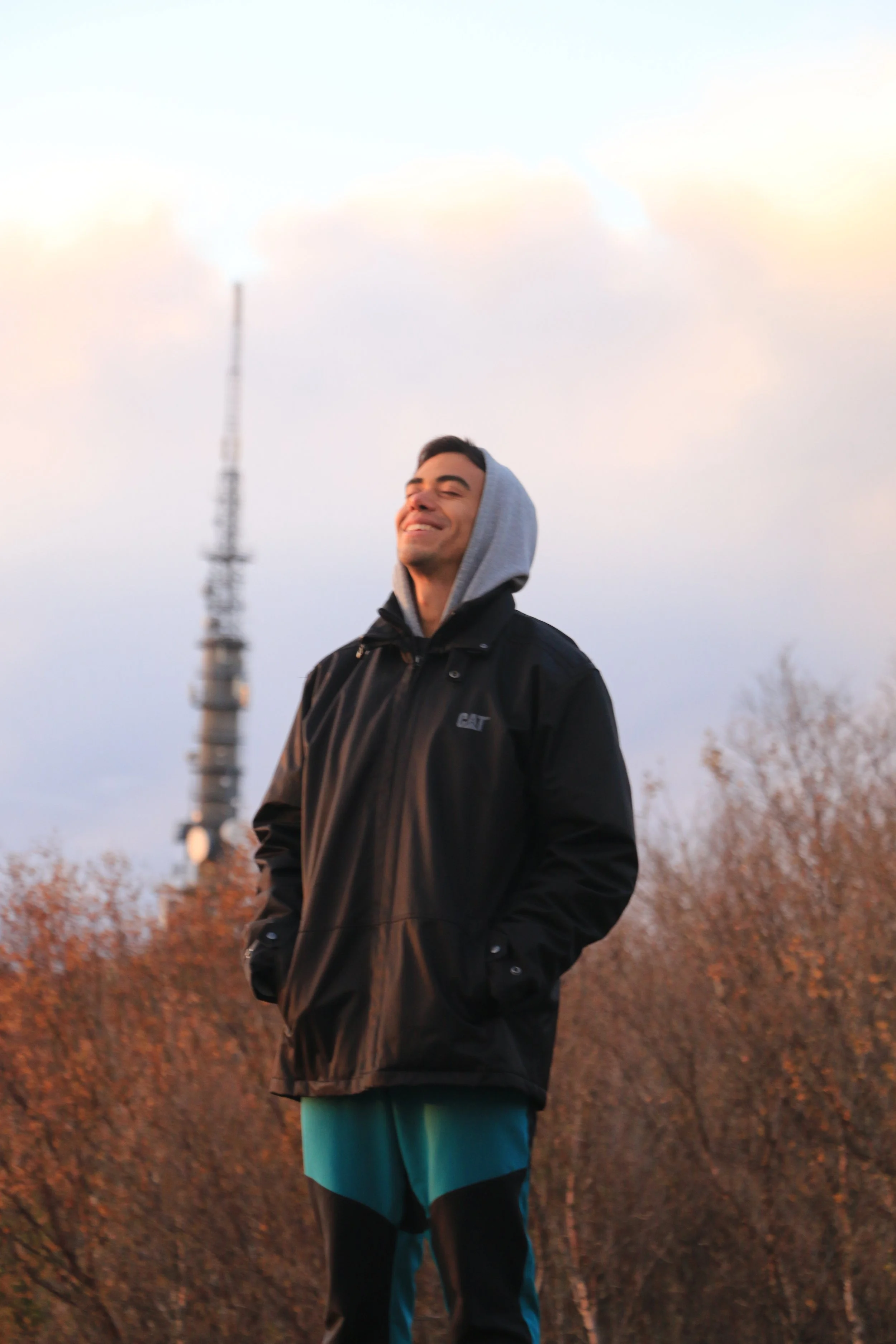 A smiling young man stands outdoors at sunset, wearing a black jacket, gray hooded sweatshirt, and teal pants, with trees and a tall tower in the background.