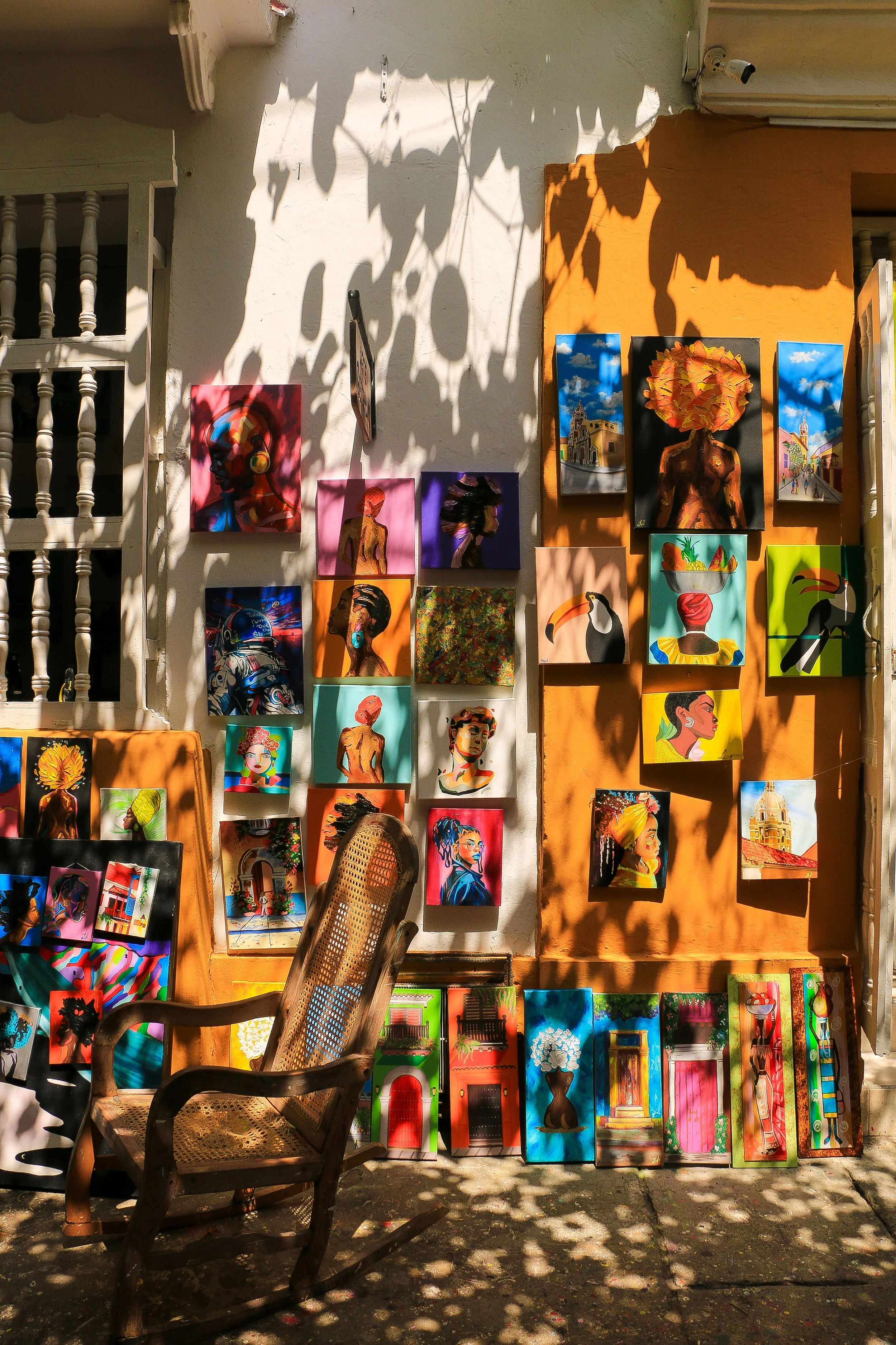 Outdoor art display with colorful paintings on a white and orange wall, with a wooden chair in the foreground, shadows of leaves, and a security camera.