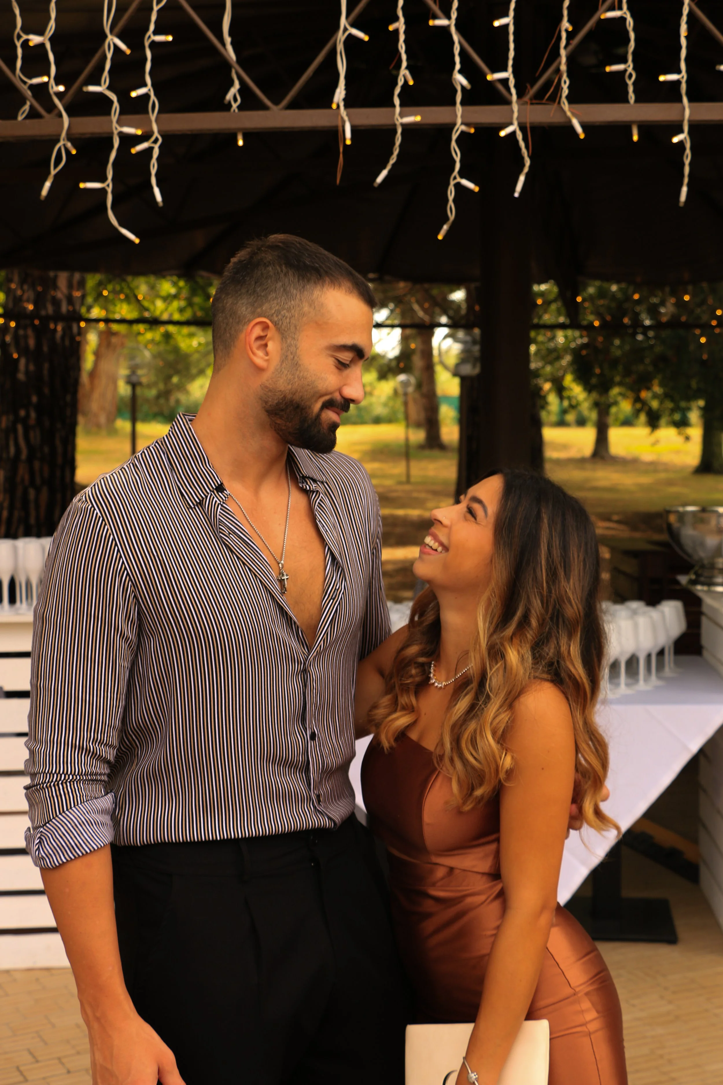 A man and woman smiling and looking at each other outdoors at a gathering or celebration, with string lights overhead and a wooded park in the background.