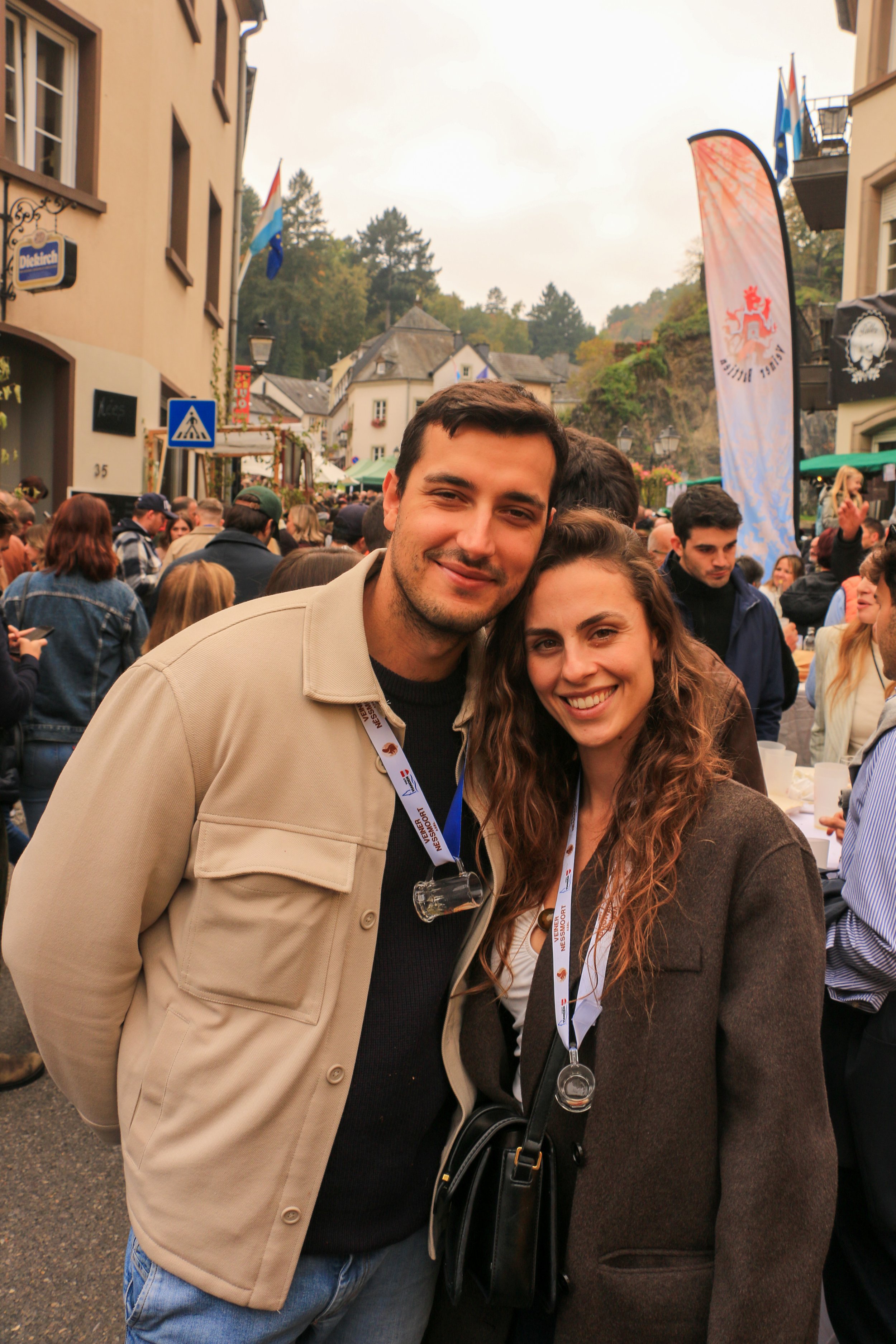 A smiling young man and woman posing together at an outdoor event with a crowd and buildings in the background.