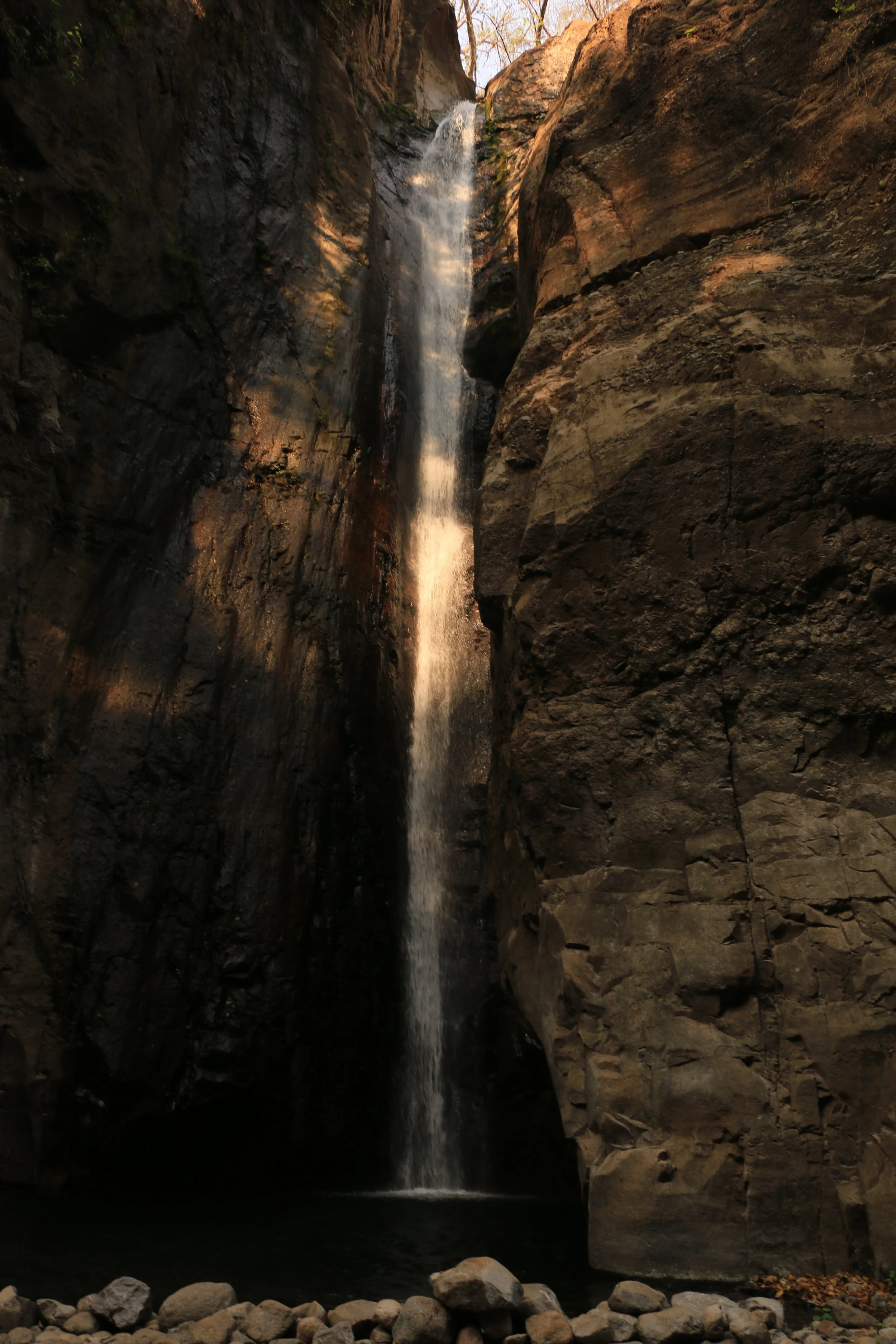 Tall waterfall flowing between rocks into a pond below, with rocky terrain at the base and some trees visible at the top.