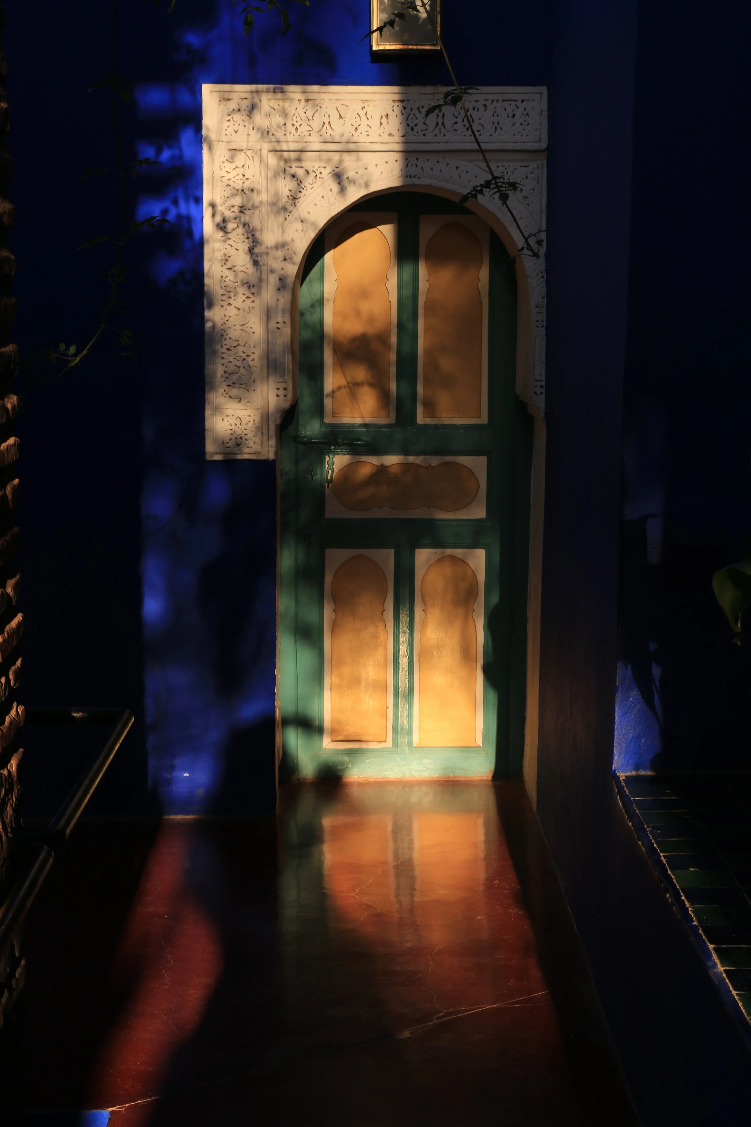 A colorful door with intricate white trim, green frame, and yellow panels, set within a deep blue wall, illuminated by warm sunlight creating shadows.