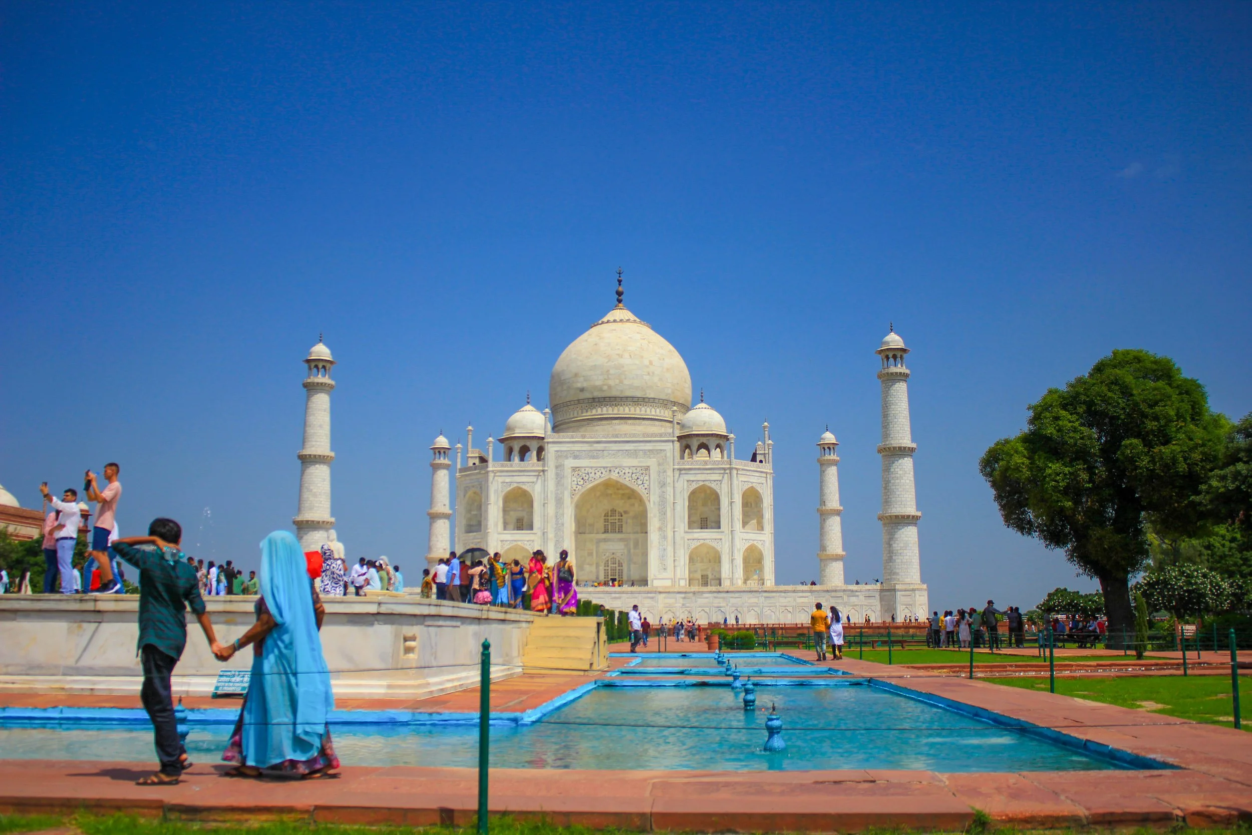 The Taj Mahal, a white marble mausoleum with a large central dome and four minarets, surrounded by visitors and gardens under a clear blue sky.