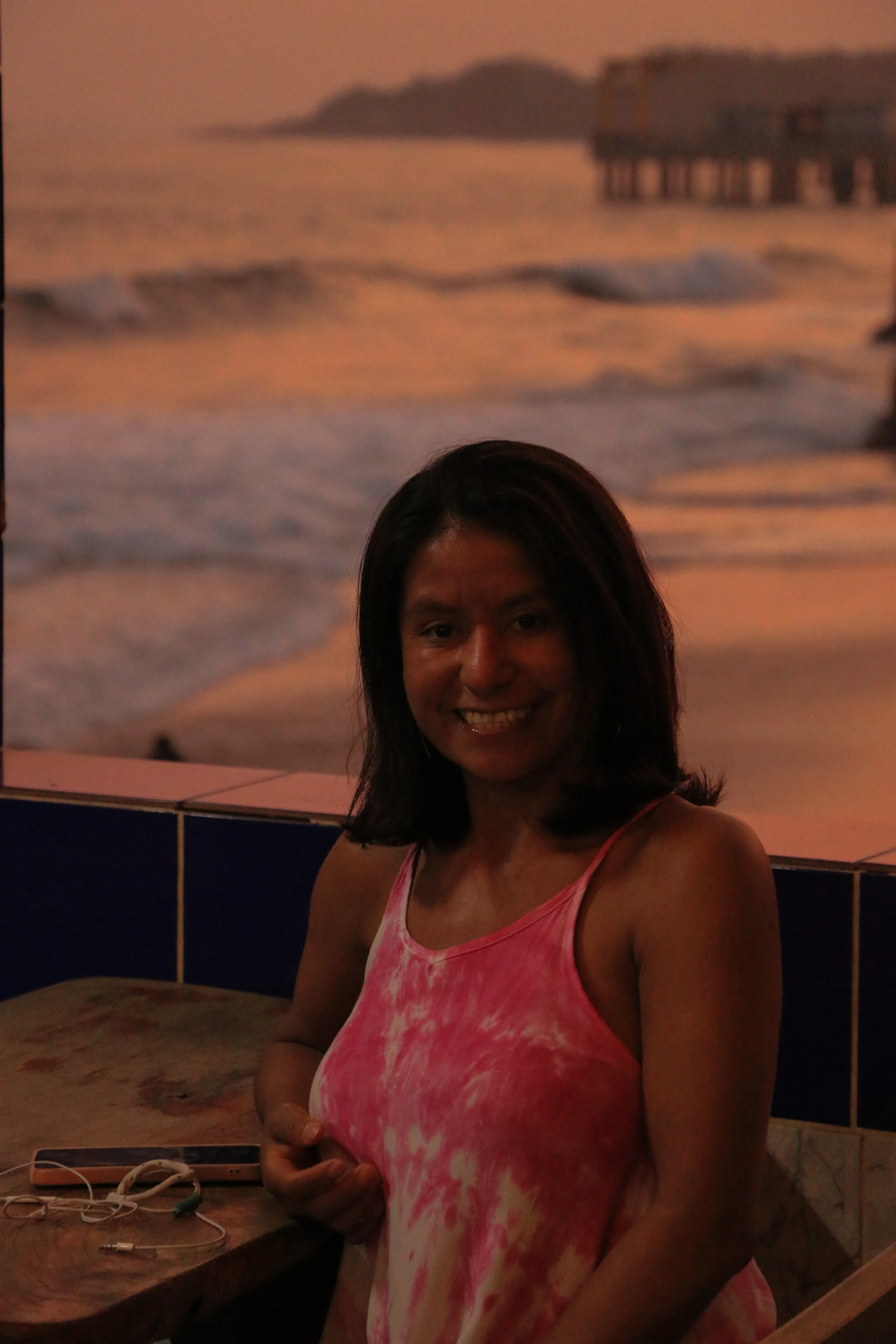 A woman standing inside near a beach-themed mural with a woman in a pink tie-dye dress smiling and holding her hand on her chest, with a table holding a smartphone and earphones.