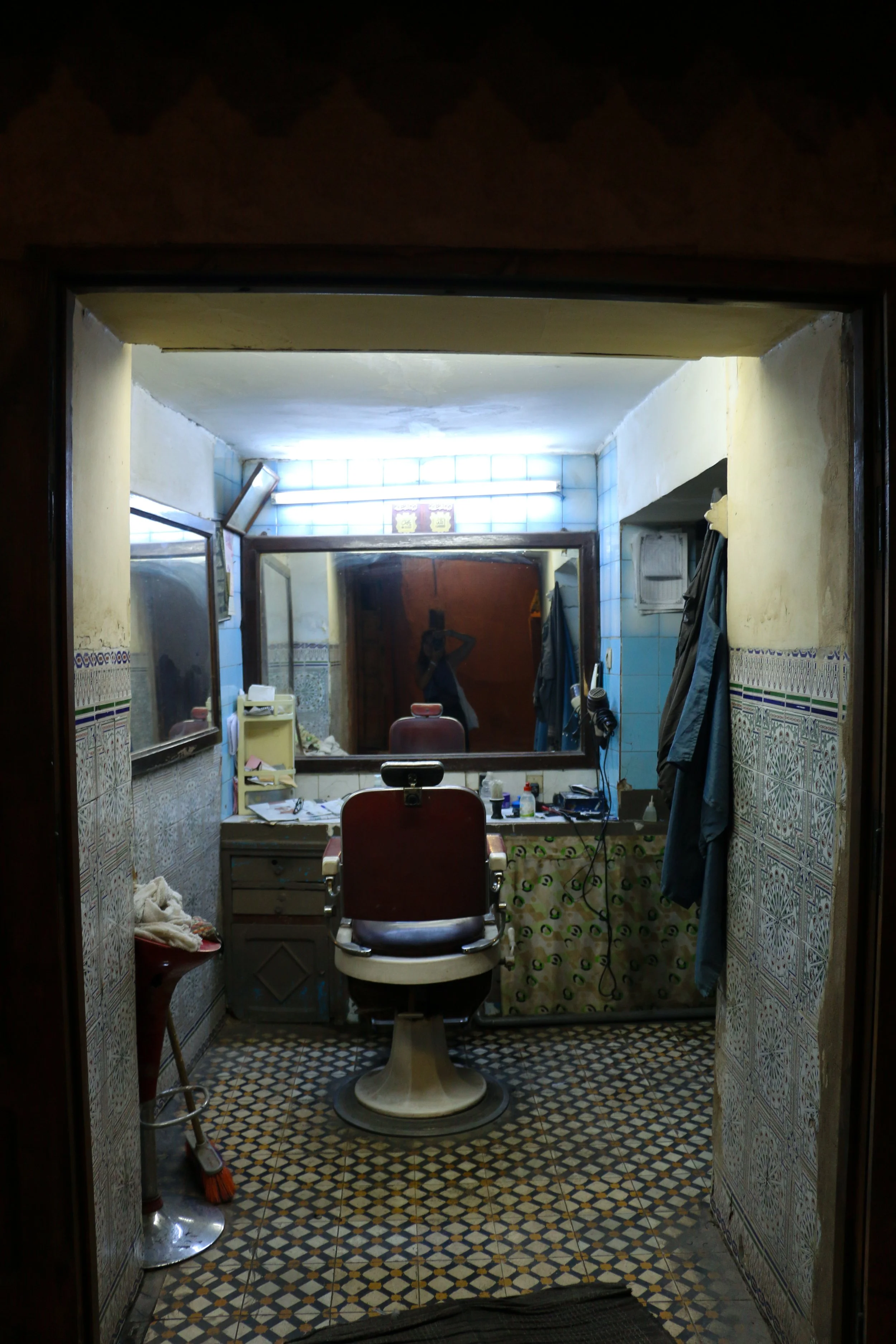 A cluttered barber shop with a barber chair in the center, a large mirror on the wall behind it, and various barber tools and supplies on the countertop. There are clothes hanging on hooks and a phone on the wall.