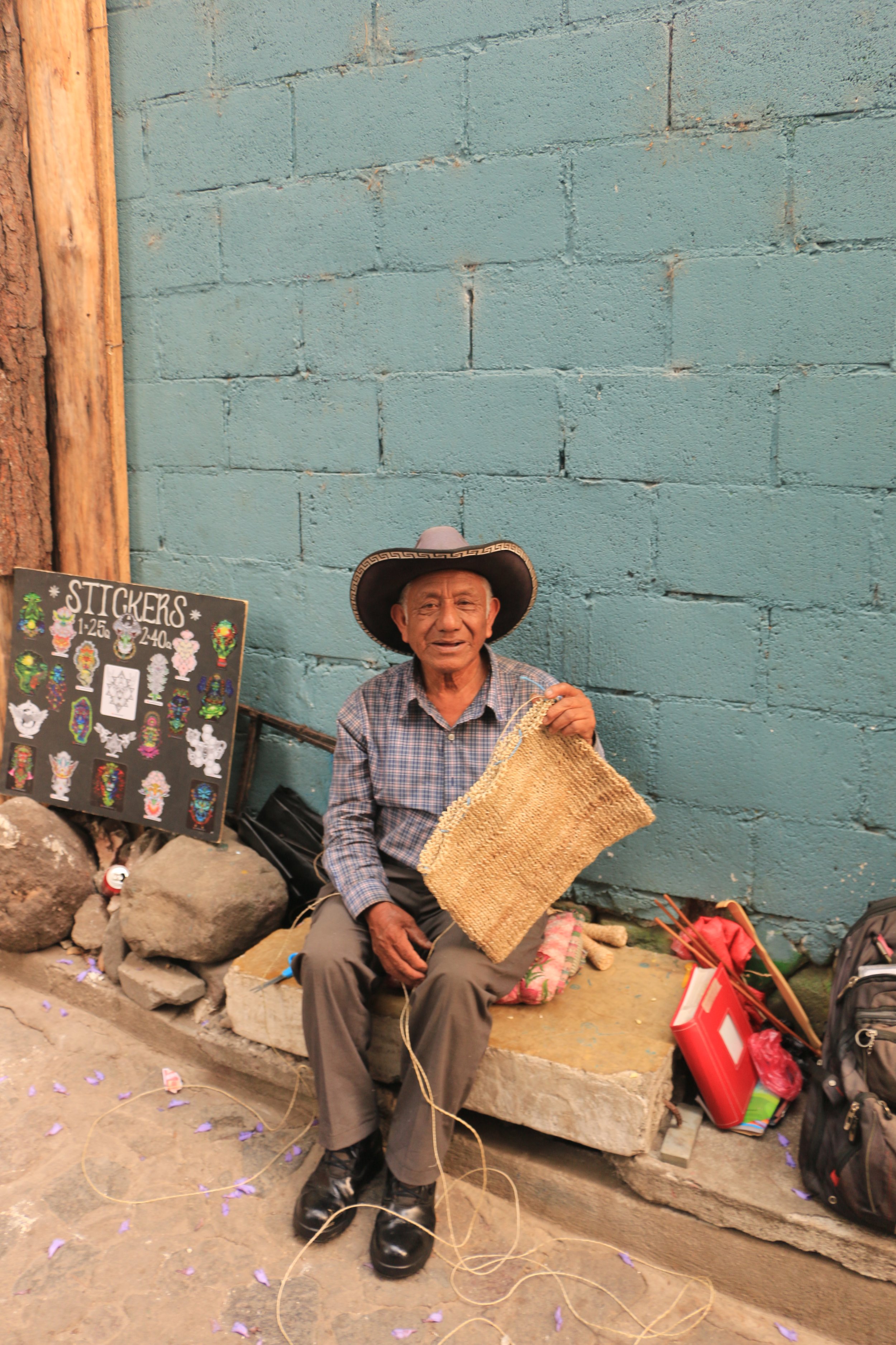 An elderly man wearing a sombrero, plaid shirt, and gray pants, sitting on a stone bench against a blue brick wall. He is holding a woven craft and smiling. Nearby are rocks, a sign with sticker designs, a backpack, and other small items.