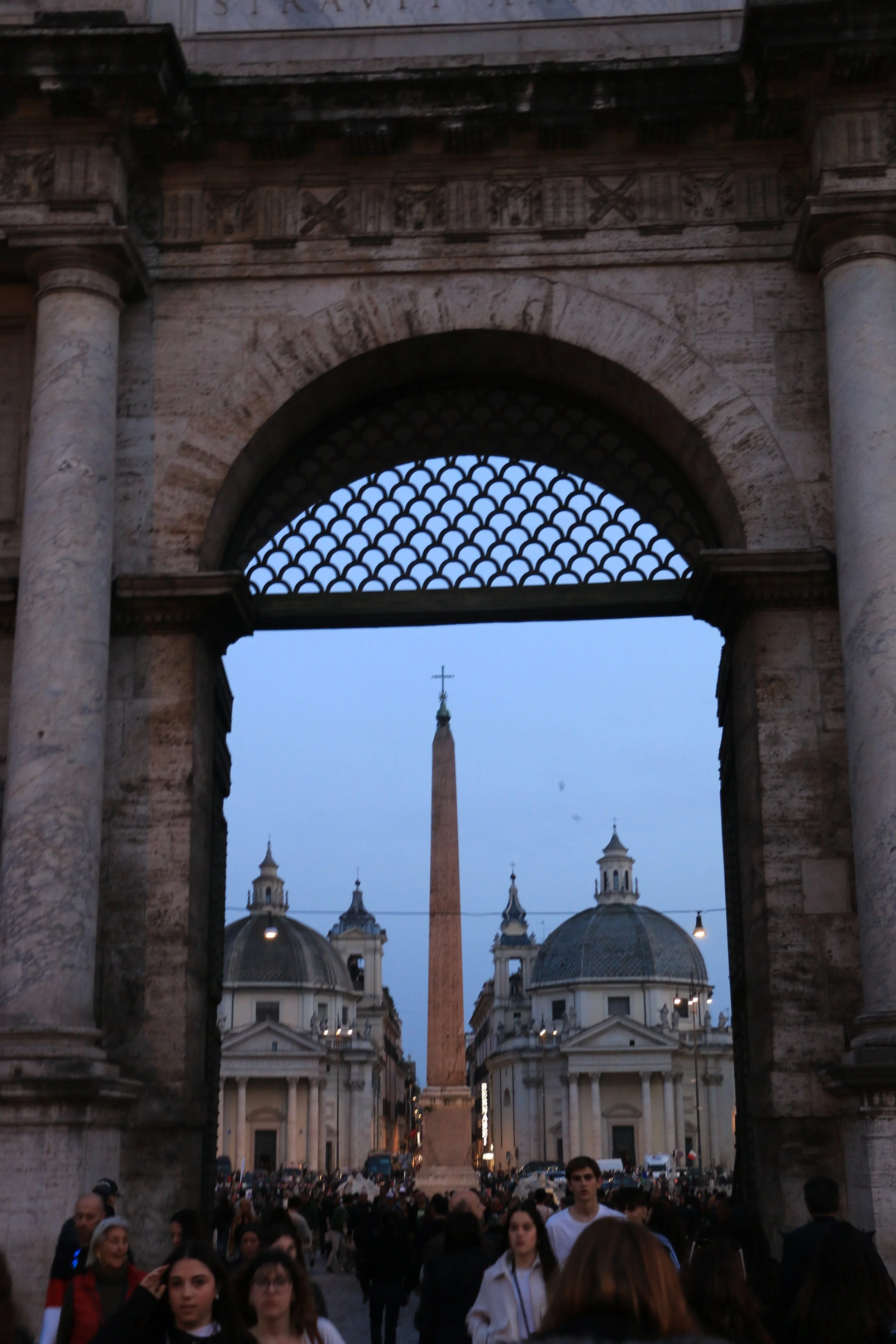 View of Piazza del Popolo in Rome, Italy, seen through an ornate archway; tourists walking, historic church buildings, an ancient Egyptian obelisk in the center, and evening sky.