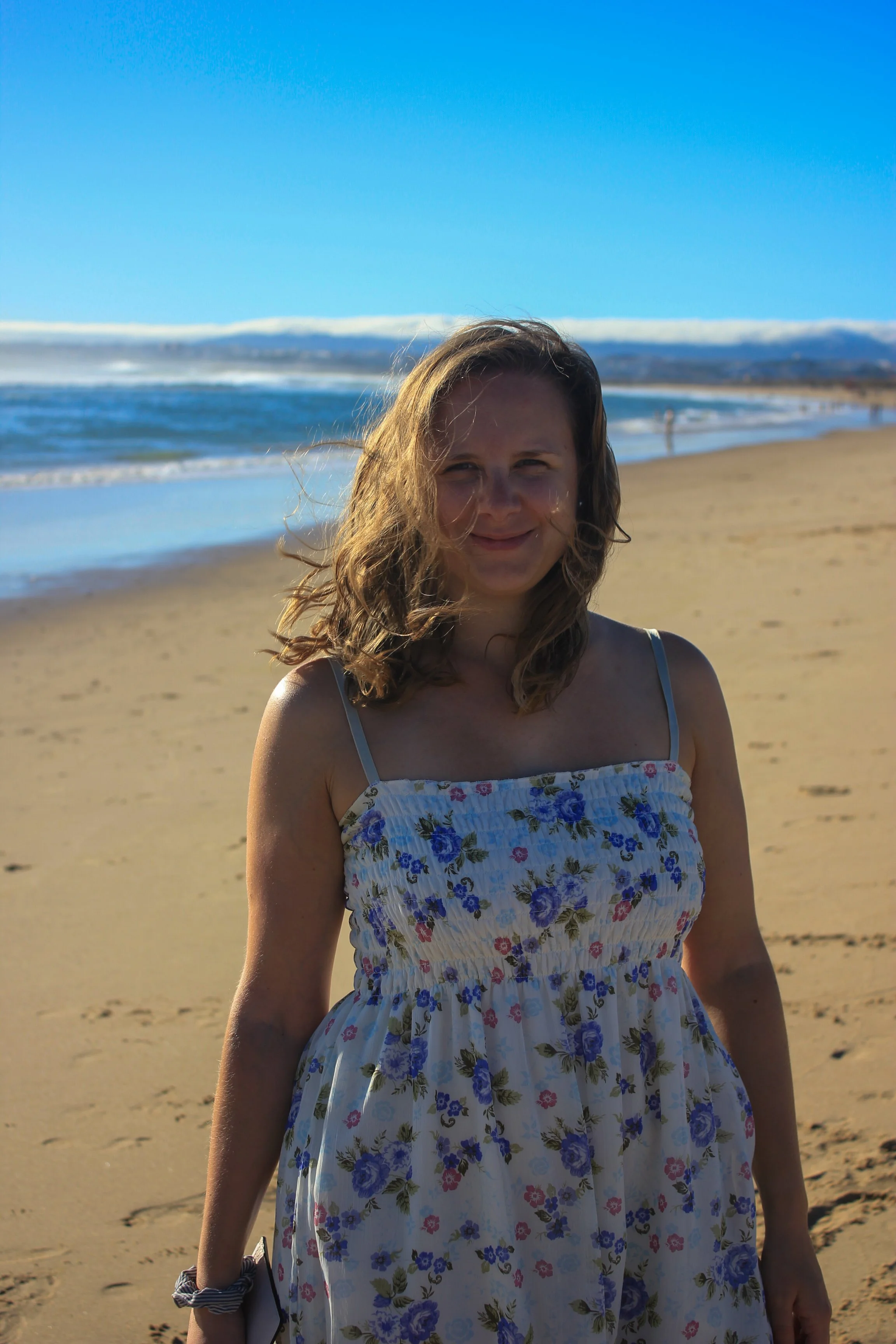 Young woman with wavy hair smiling on a sandy beach with ocean and blue sky in the background.