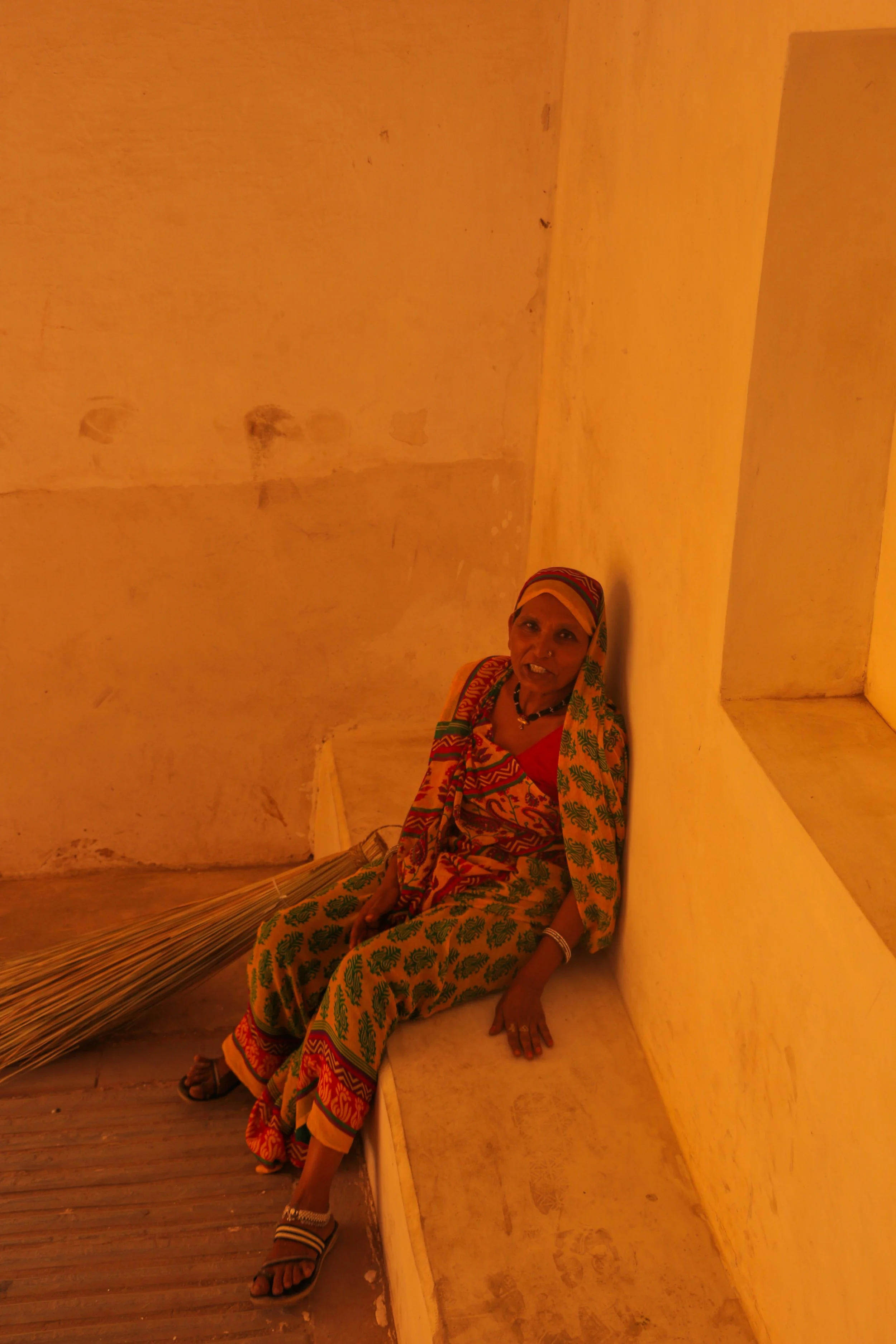 An elderly woman with dark skin, wearing traditional colorful clothing and a headscarf, sitting on a bench in a room with yellowish lighting and plain walls.