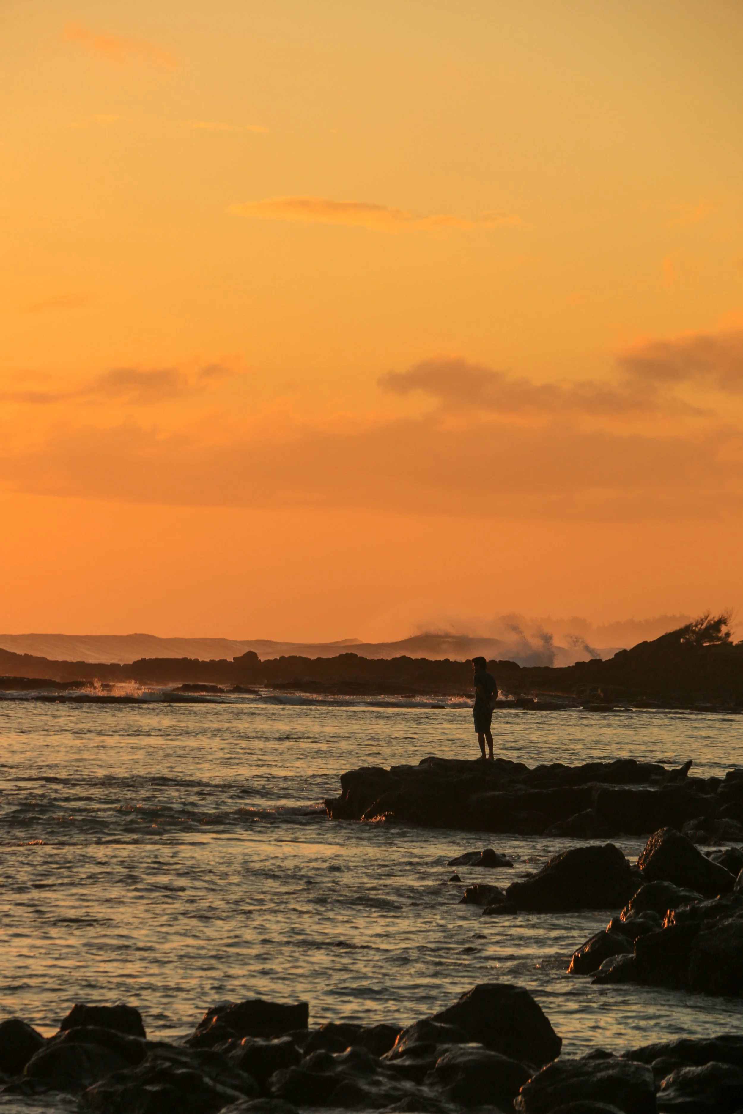 Person standing on rocks by the ocean at sunset with a colorful sky.