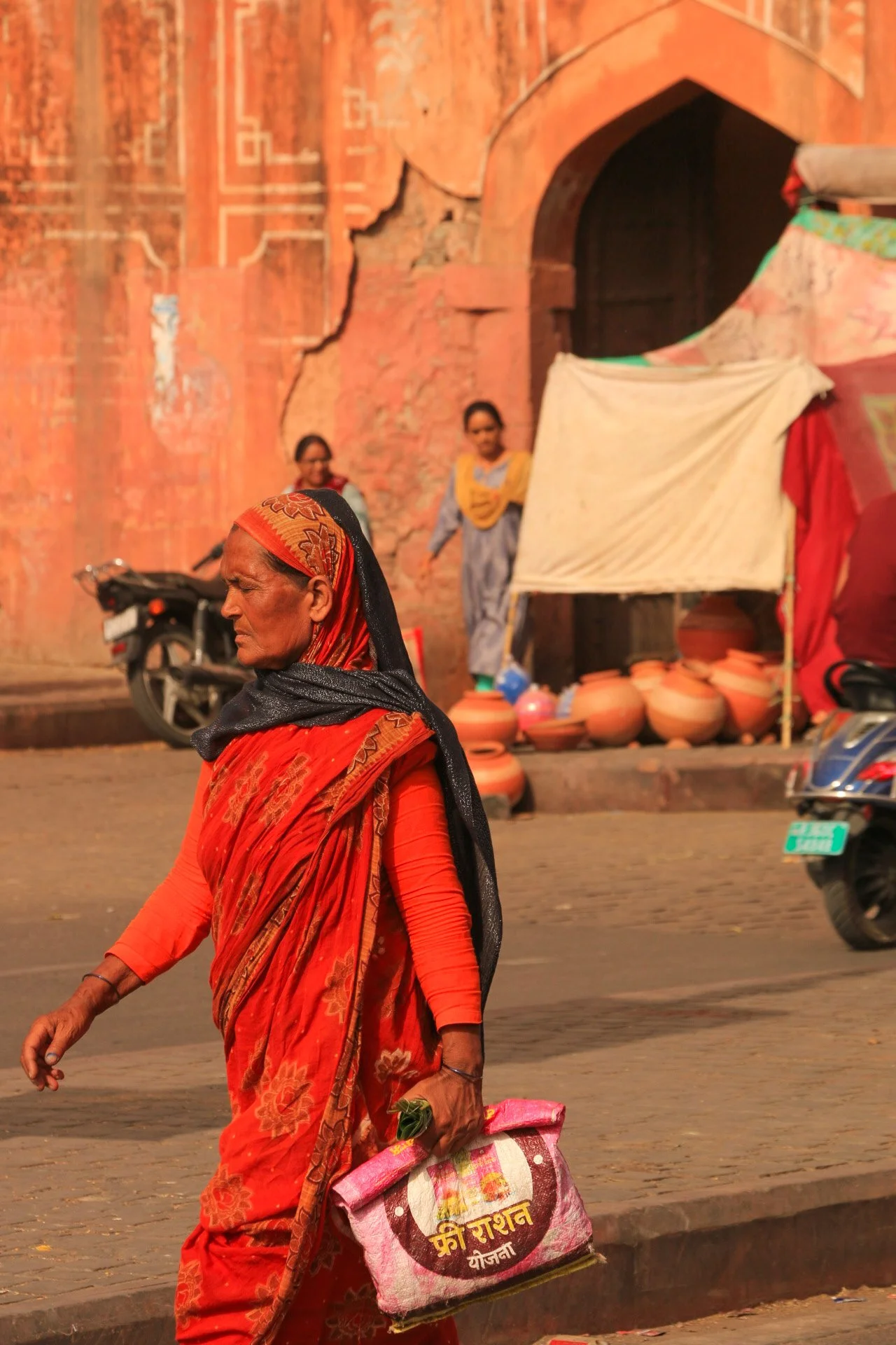 An elderly woman dressed in a red sari and headscarf walking along a street in front of a brick wall. She is carrying a pink bag with yellow and red writing on it, and she is holding a small green object in her right hand.