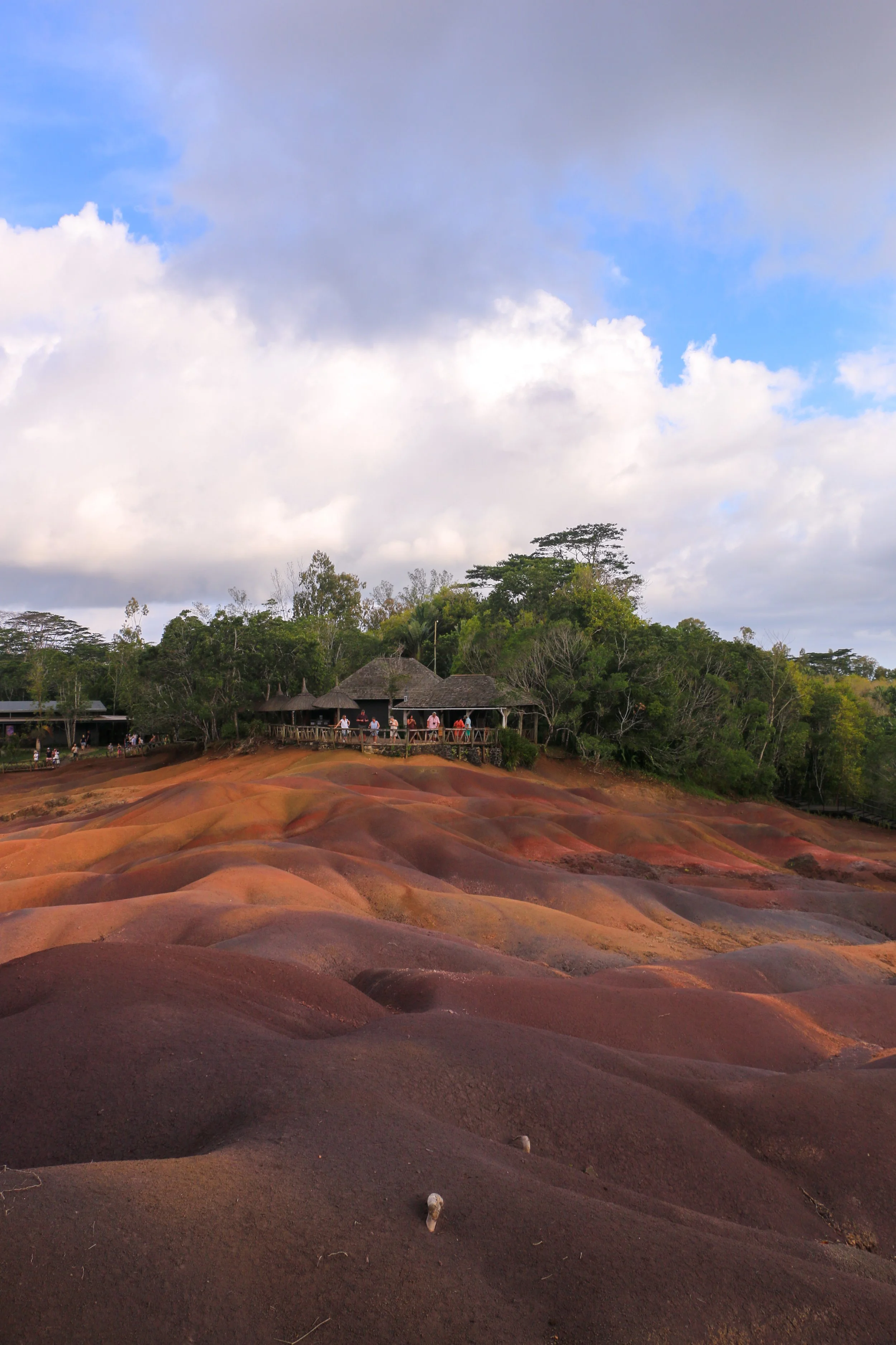 Colorful rolling hills with reddish-brown and purple hues, a wooden structure with people on a balcony, and a green forest in the background under a partly cloudy sky.