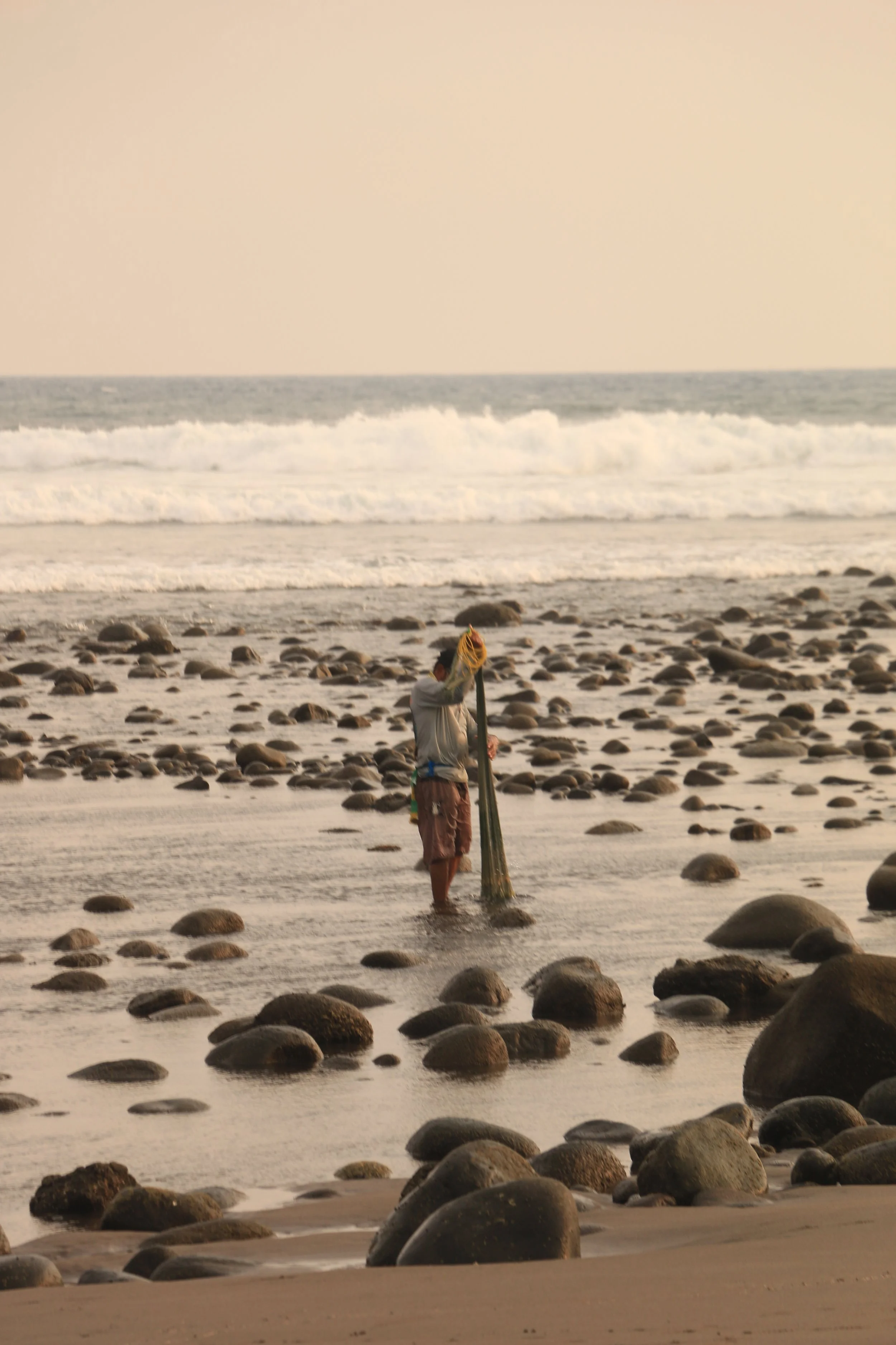 A person standing in shallow water on a rocky beach, holding a long pole and looking down at it, with ocean waves in the background.